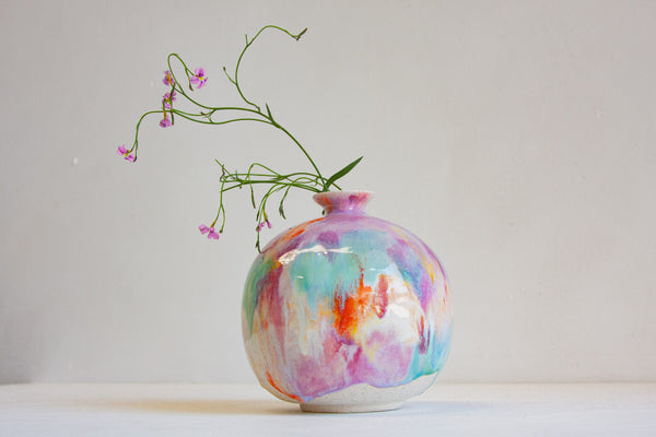 A round ceramic vase with a colorful marbled pattern in pink, purple, blue, and orange, holding delicate green stems with small pink flowers, set against a plain light gray background.