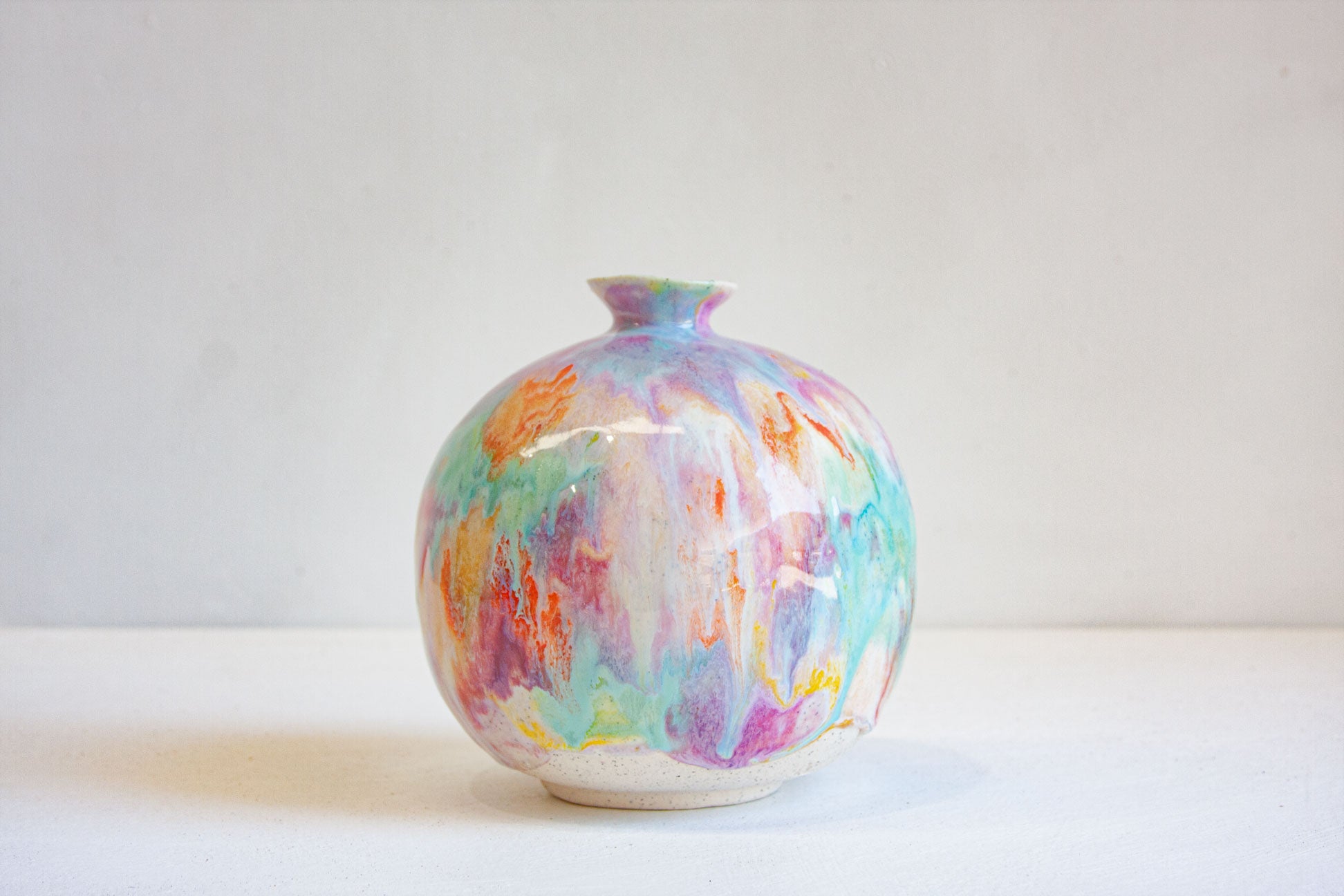 A small round ceramic vase with a narrow neck, decorated with a colorful marbled glaze in pastel shades of purple, blue, pink, orange, and yellow, set against a plain light gray background.