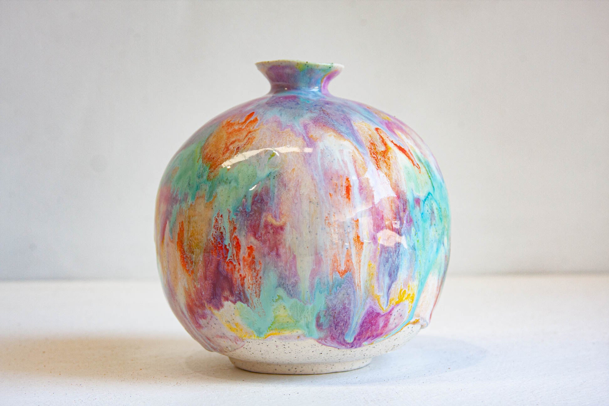 A round ceramic vase with a glossy finish, decorated with colorful abstract marbled paint patterns in purple, blue, green, orange, and pink on a white base, set against a plain light gray background.