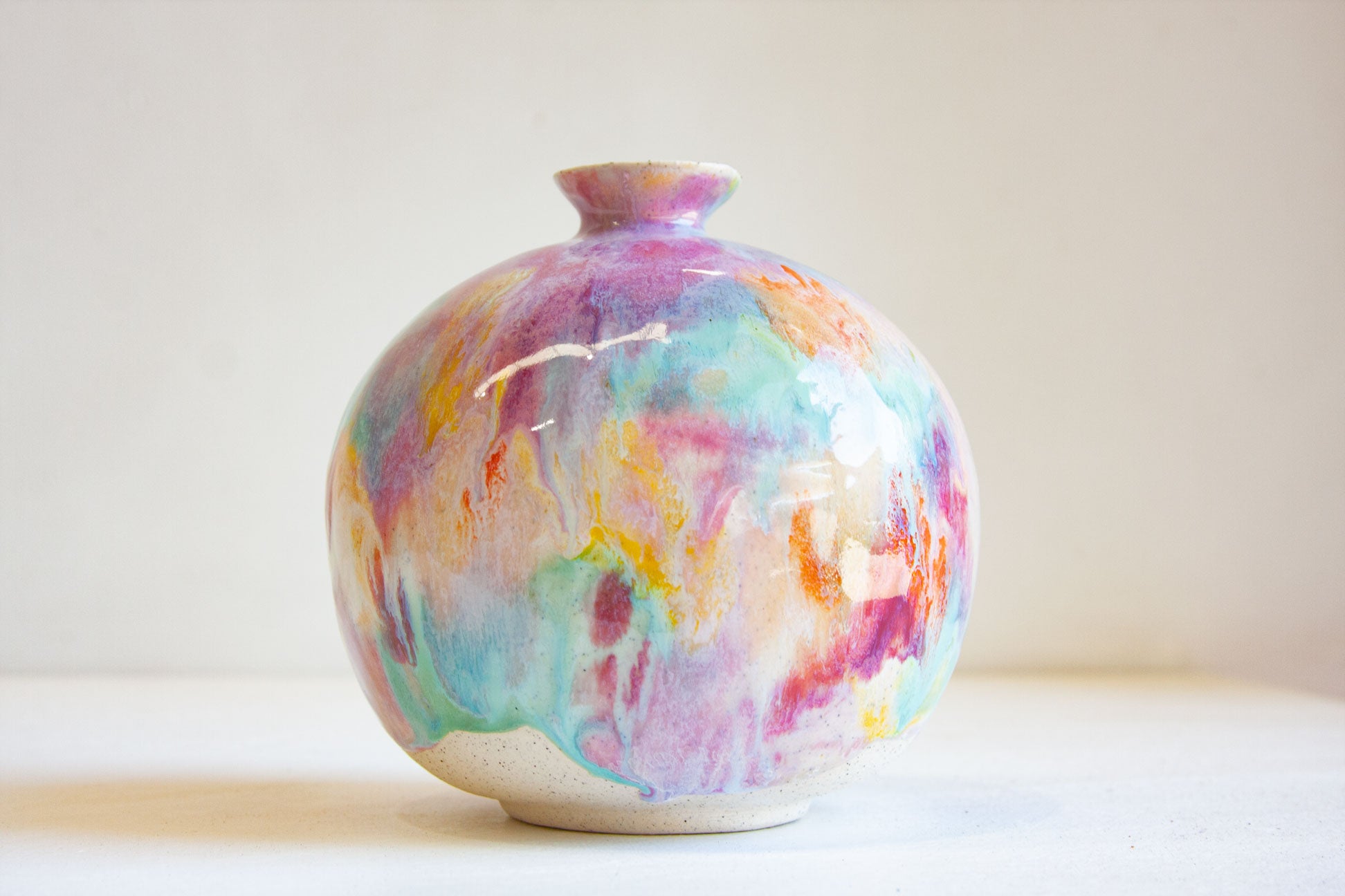 A small round ceramic vase with a short neck, decorated with a colorful marbled glaze in shades of pink, purple, blue, teal, and orange, set against a plain light beige background.