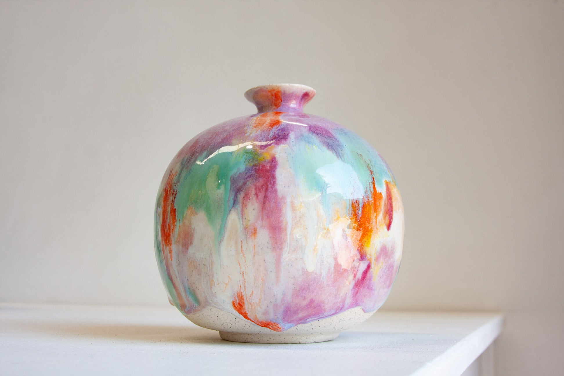 A round ceramic vase with a glossy finish, decorated with a colorful abstract drip pattern in pink, purple, teal, orange, and white, displayed on a white shelf against a neutral beige background.