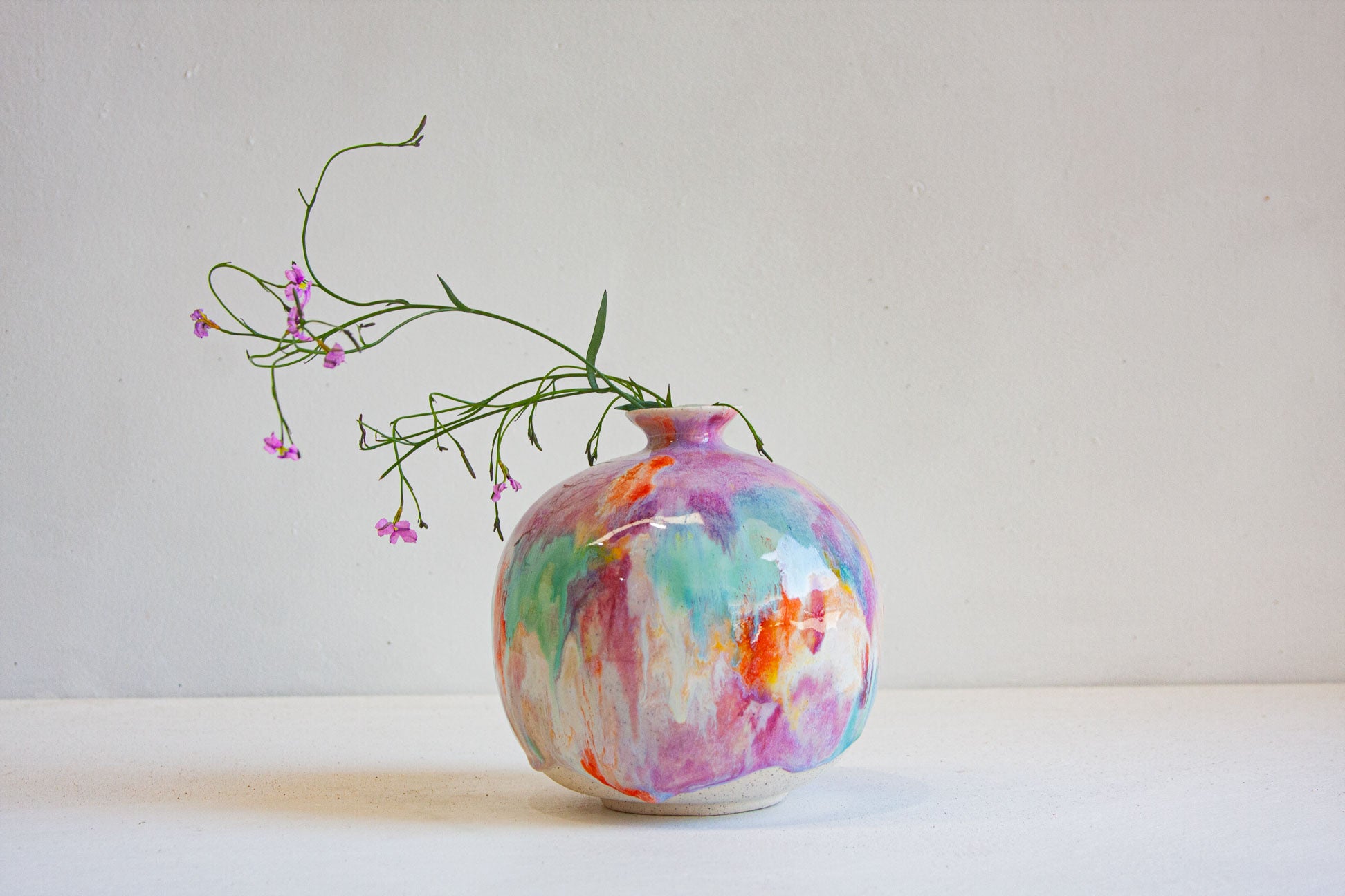A small round ceramic vase with a glossy finish, decorated in a colorful abstract marbled pattern of pink, purple, blue, green, and orange on a white base. A delicate green sprig with tiny pink flowers is placed in the vase, set against a plain off-white background.