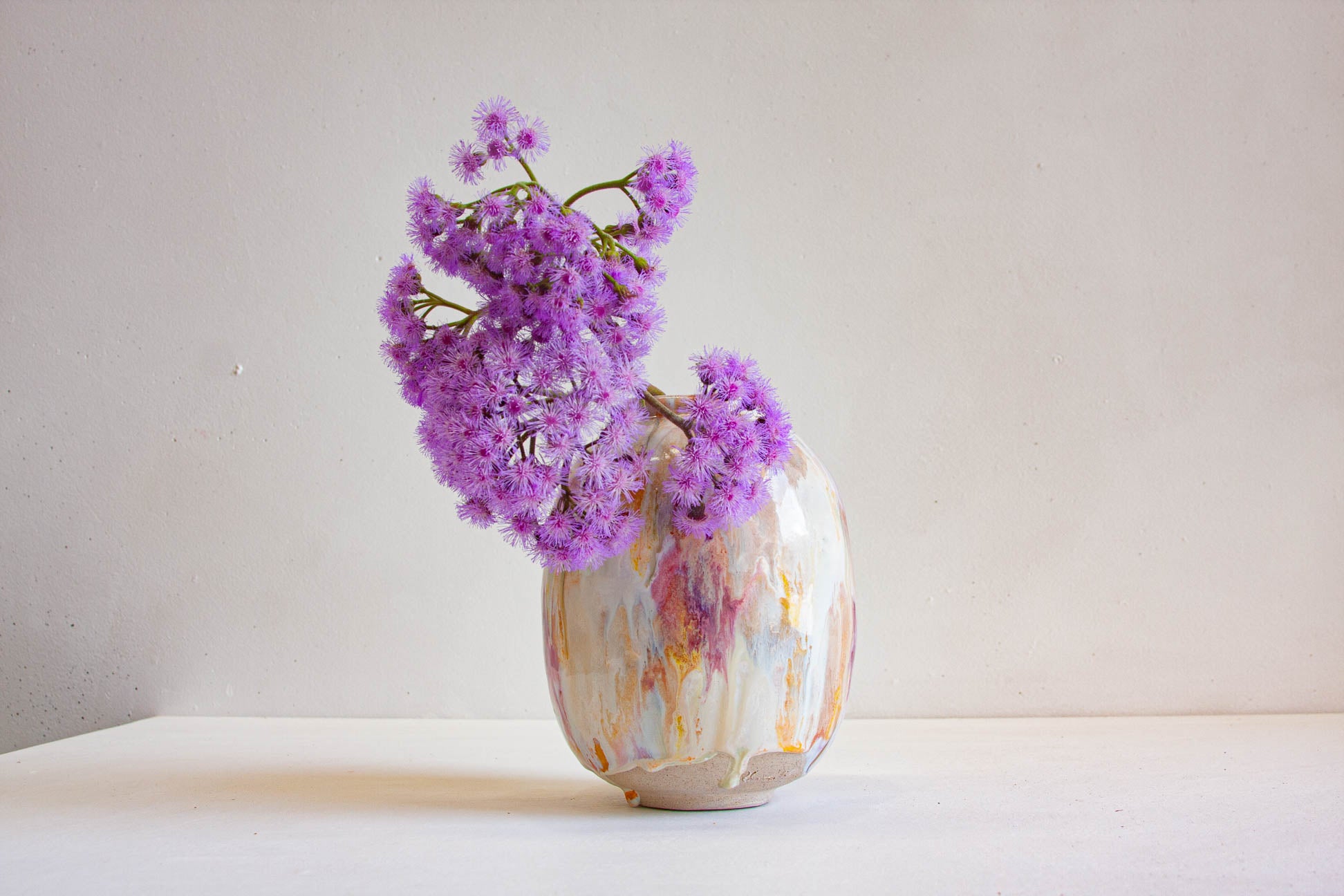 A small rounded ceramic vase with a marbled off-white, pink, and gold glaze, holding a cluster of purple flowers, set against a plain light gray wall and white surface.