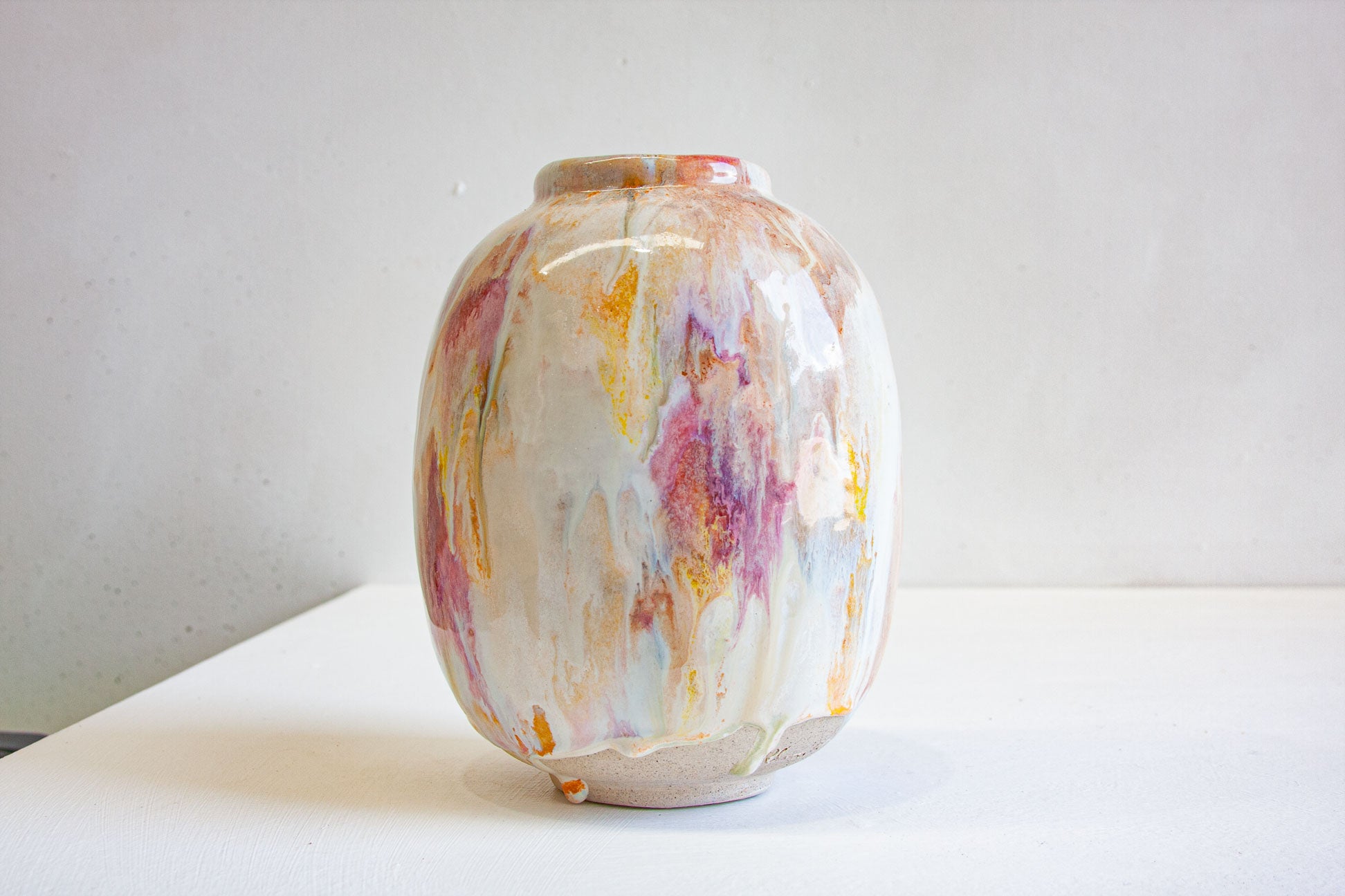 A small ceramic vase with a glossy finish, decorated with a marbled, paint-splatter pattern in white, pink, orange, and yellow, sitting on a white surface against a light gray wall.
