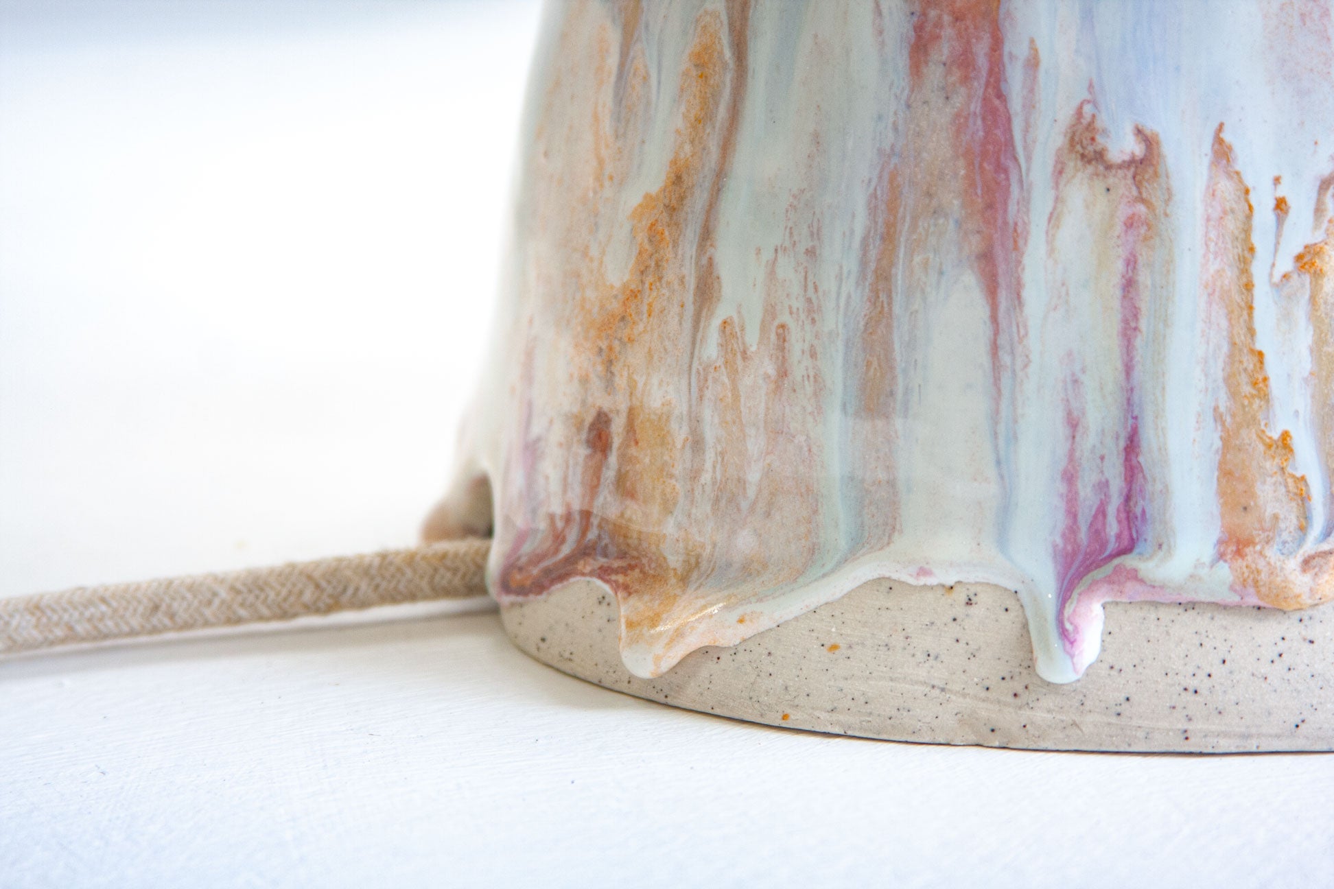 Close-up of a ceramic mug showing a marbled glaze with white, pink, and orange-brown drips over a speckled beige clay base, with a woven straw strap visible on the left.