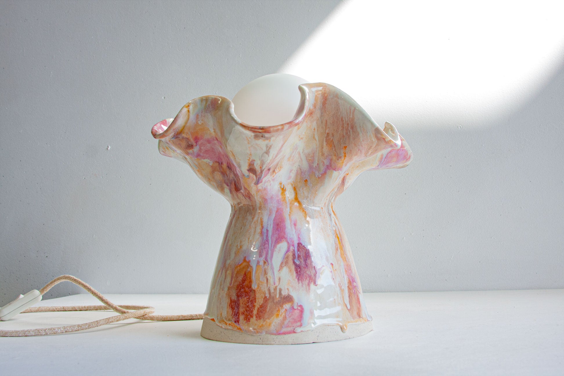 A ceramic lamp with a sculptural, draped silhouette in a glossy white glaze decorated with pink, orange, and brown marbled patterns. A warm white globe bulb is inserted into the top opening, and a beige power cord lies on the white surface to the left.