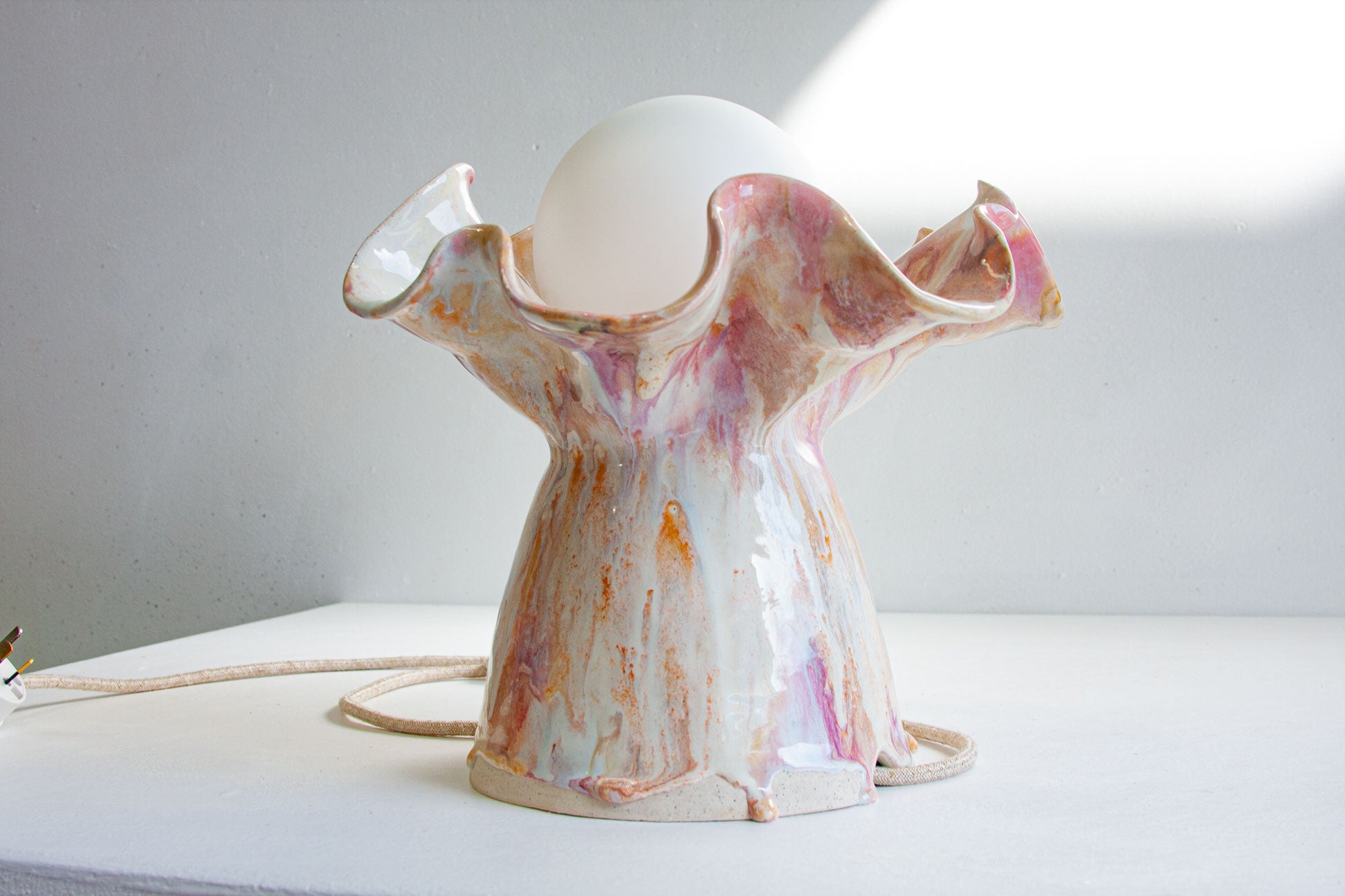 A sculptural ceramic table lamp with a glossy, marbled finish in white, pink, and orange, featuring a flared rim and a white globe shade, shown on a white surface against a gray wall.