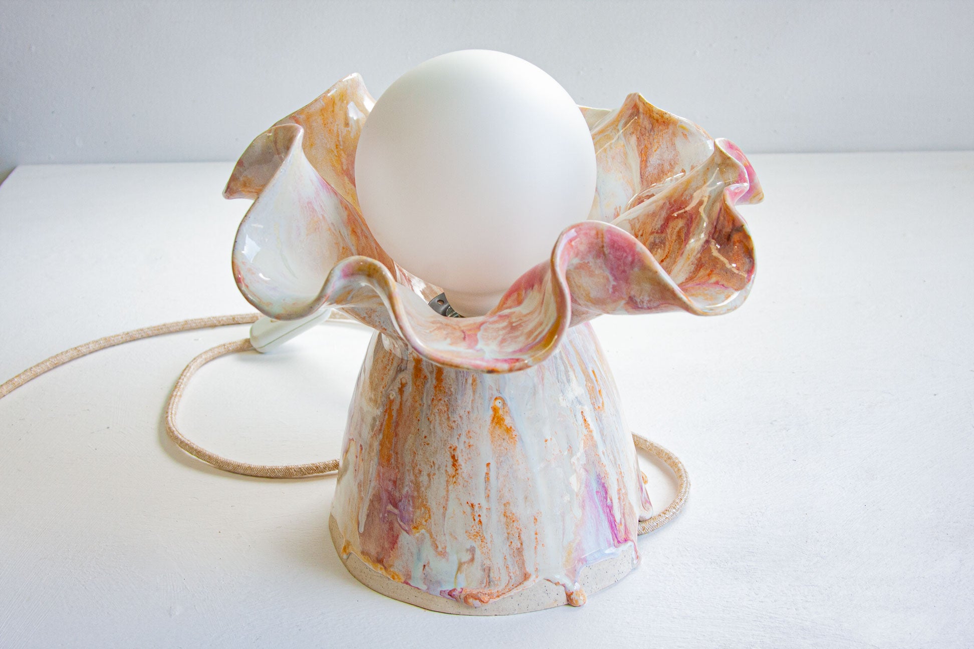 A sculptural ceramic lamp base with a white globe bulb inserted, featuring a marbled glaze in cream, pink, and orange tones, with a coiled cord on a white surface.