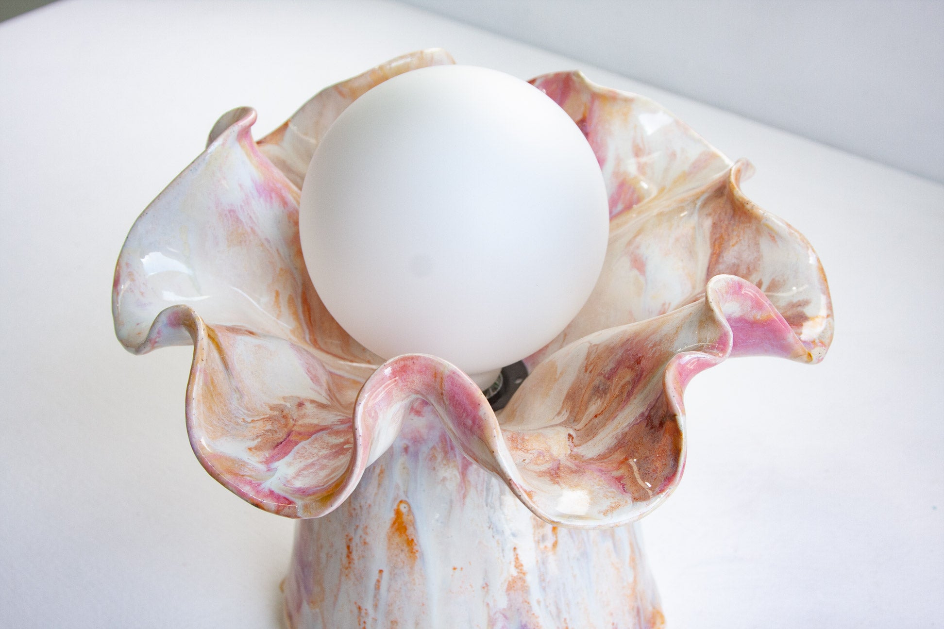 A white spherical light bulb nestled inside a sculptural ceramic holder with ruffled petals in white, pink, and brown marbled glaze, set against a plain white background.