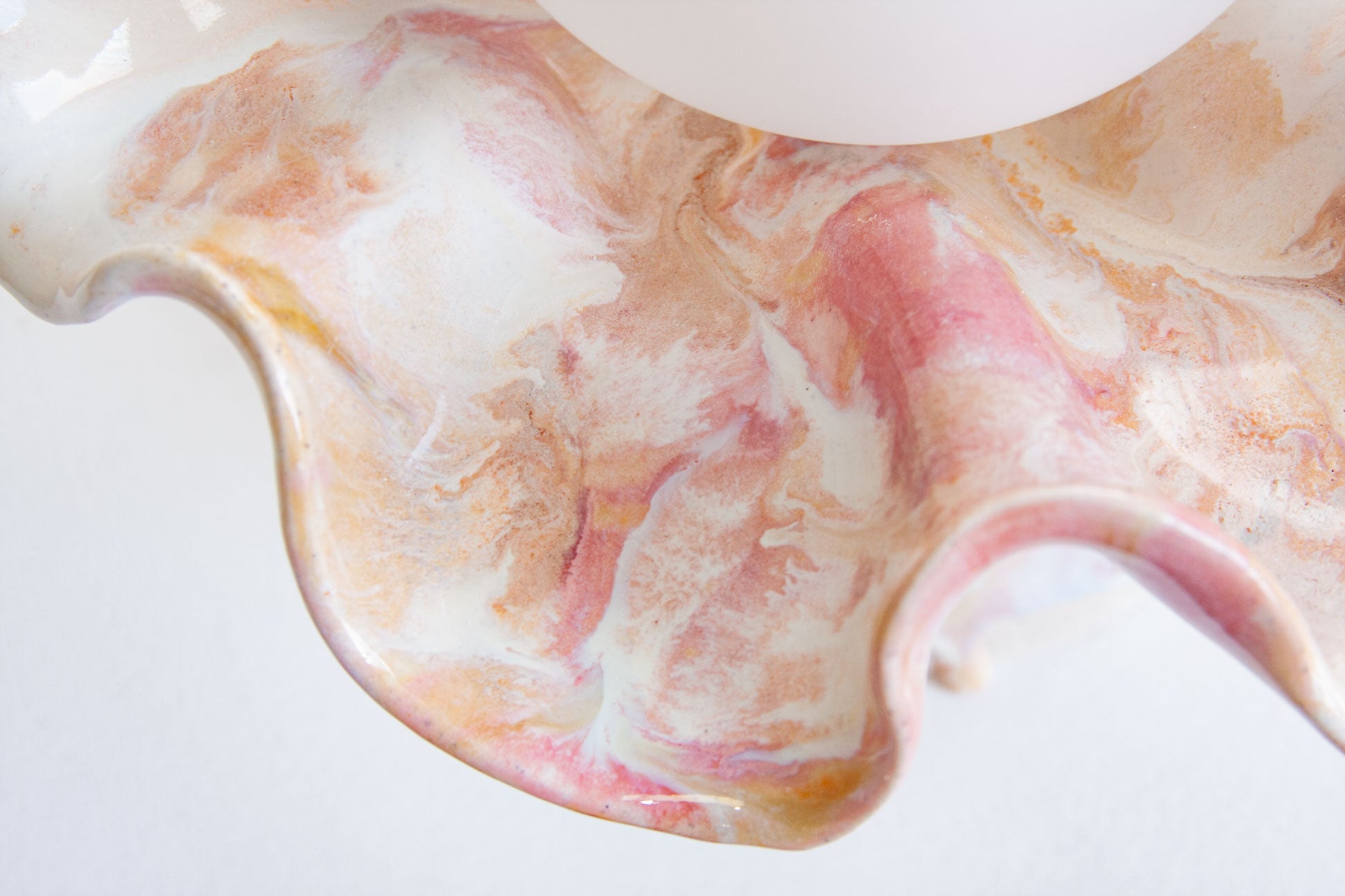 A close-up of a glossy ceramic bowl with a wavy rim, featuring a marbled pattern of white, pink, and peach-orange swirls on a white background.