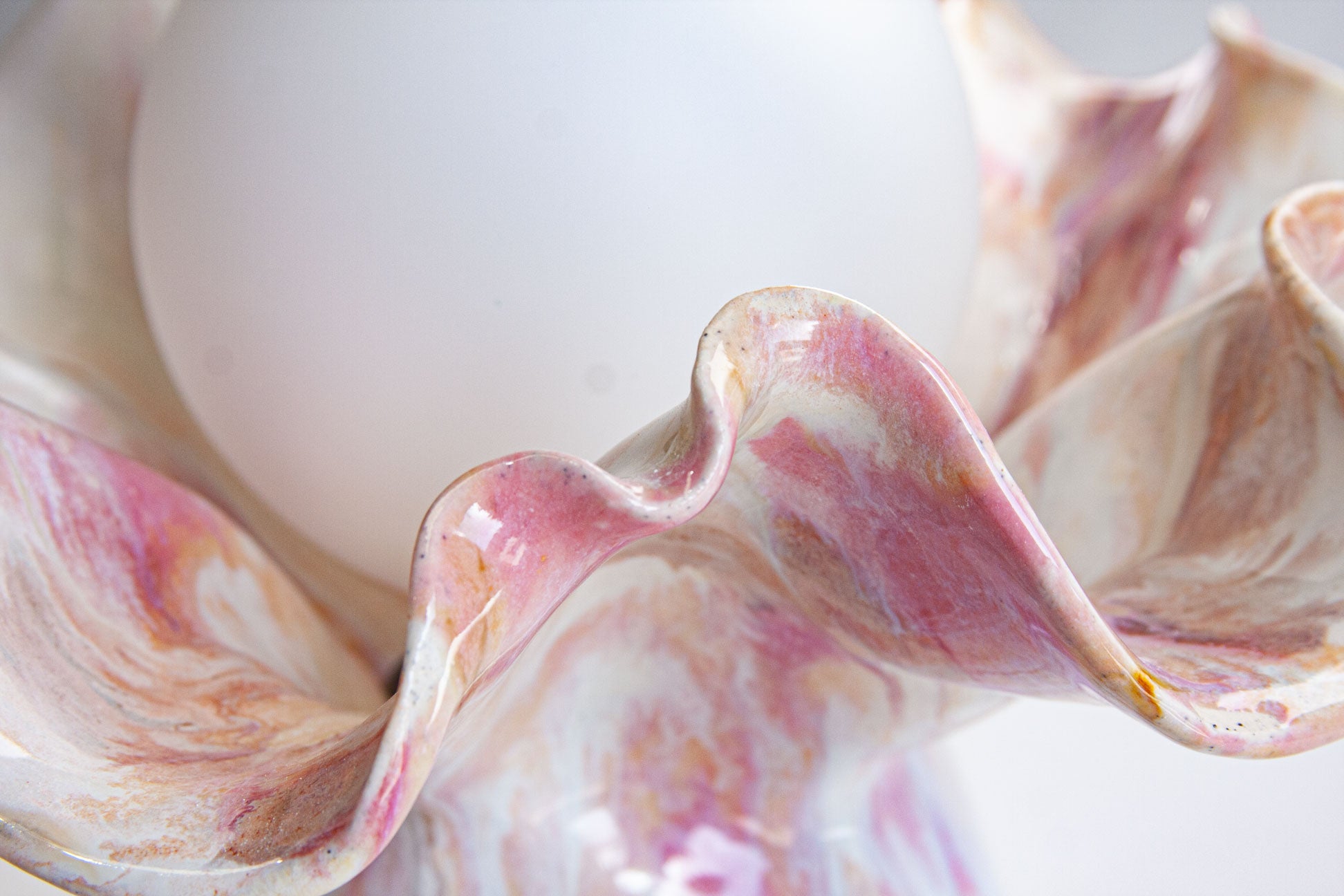 A close-up of a sculptural ceramic shell holder with fluted, clam-like ripples in pink, white, and beige tones, holding a smooth white egg.
