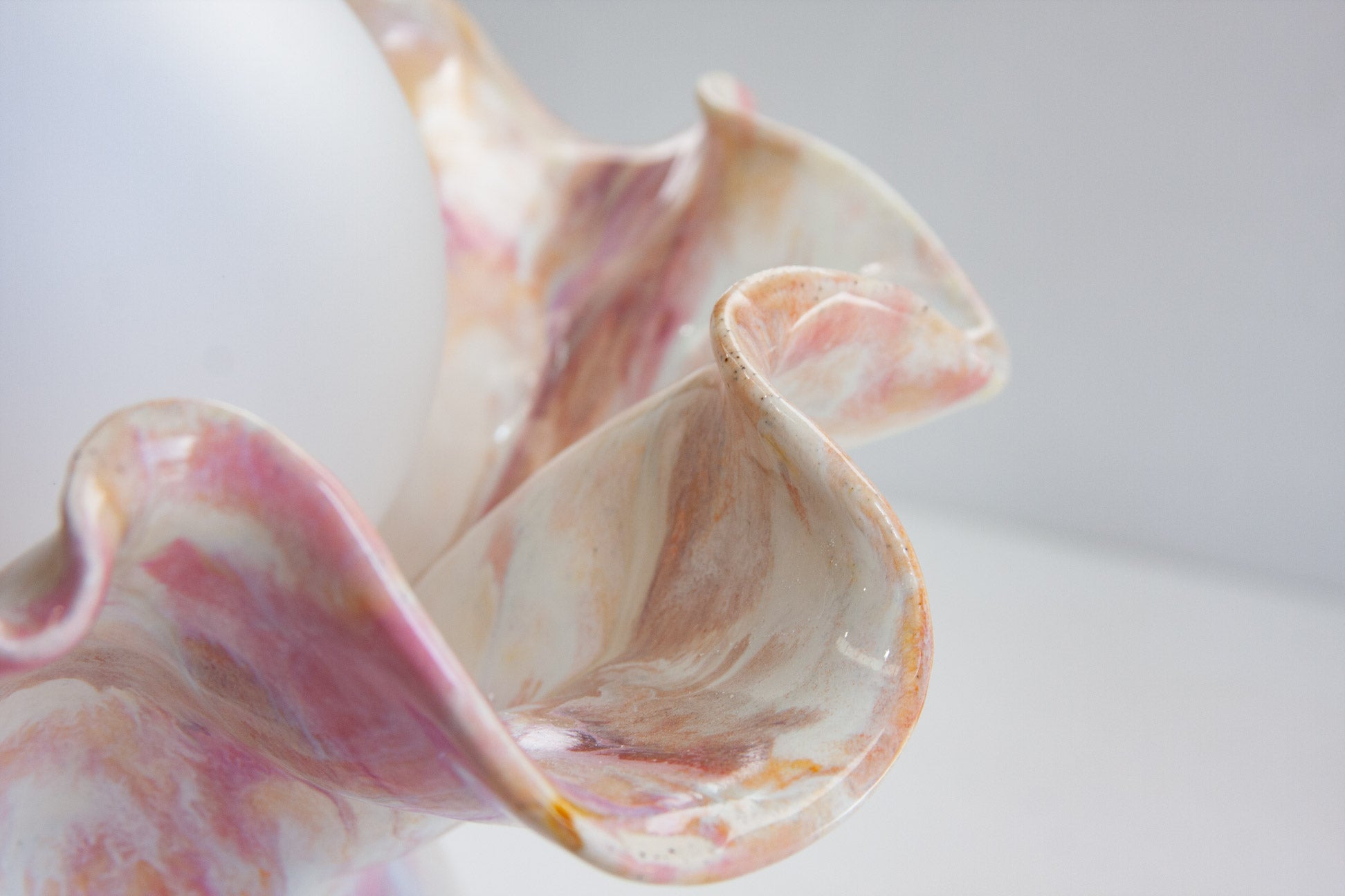 A close-up of a sculptural ceramic vessel with flared, shell-like ruffled edges in soft pink, cream, and warm brown marbled tones against a light gray background.