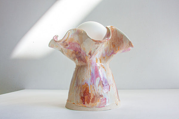 A sculptural ceramic lamp holder with a flared, ruffled top and a bulb-shaped base, featuring a marbled glaze in cream, pink, peach, and orange tones, set against a minimalist white background with a diagonal light beam.