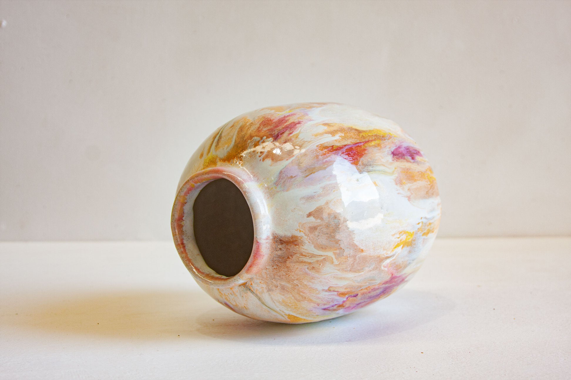 A small hand-painted ceramic vase with a marbled, fluid-art pattern in white, orange, pink, and yellow, photographed on a light beige surface against a neutral background.