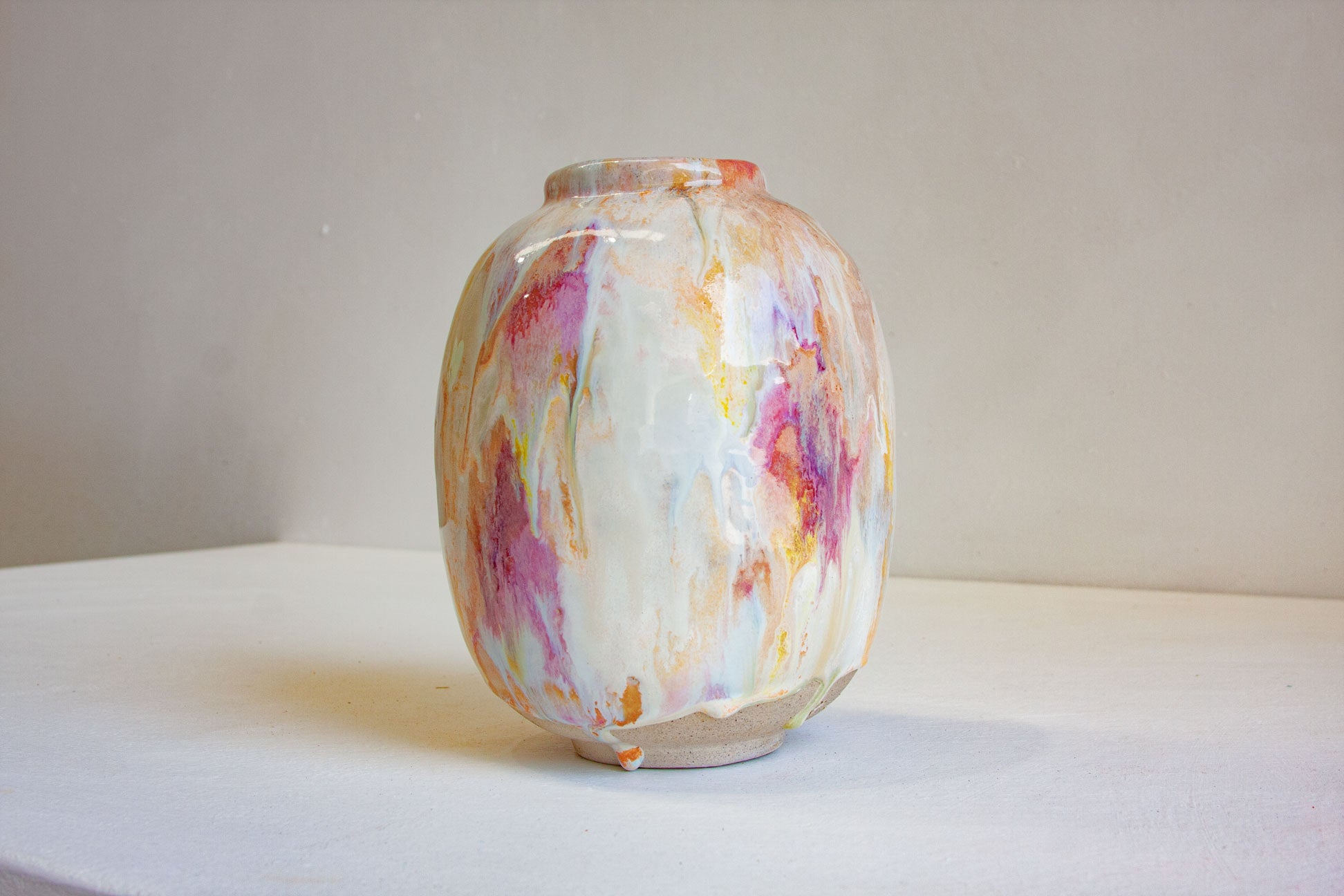 A small ceramic vase with an abstract marbled pattern in white, pink, purple, orange, and yellow, sitting on a white surface against a neutral beige background.