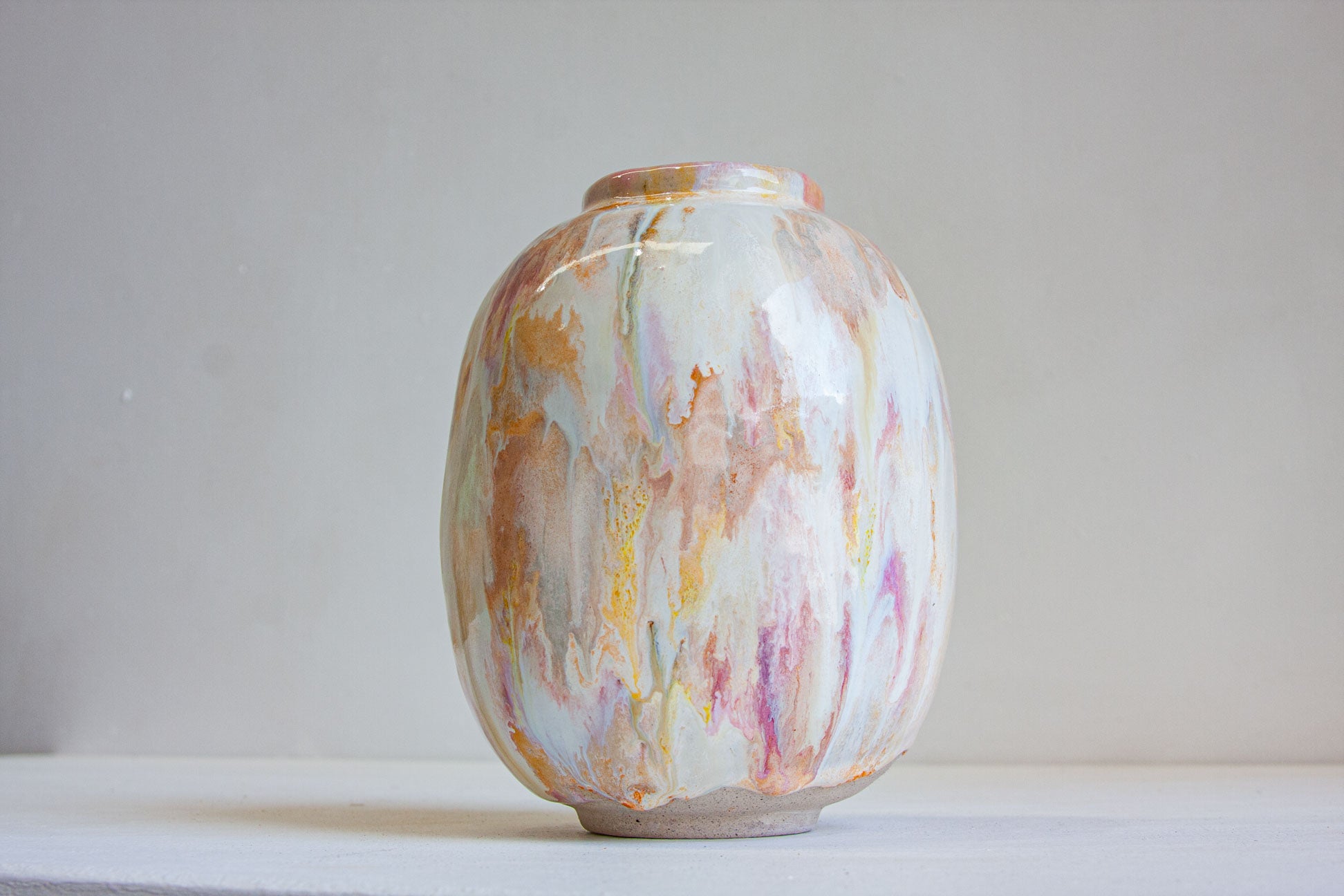 A small ceramic vase with a rounded body and short neck, decorated with a marbled, fluid-art pattern in cream, orange, pink, and yellow tones, set against a plain light gray background.