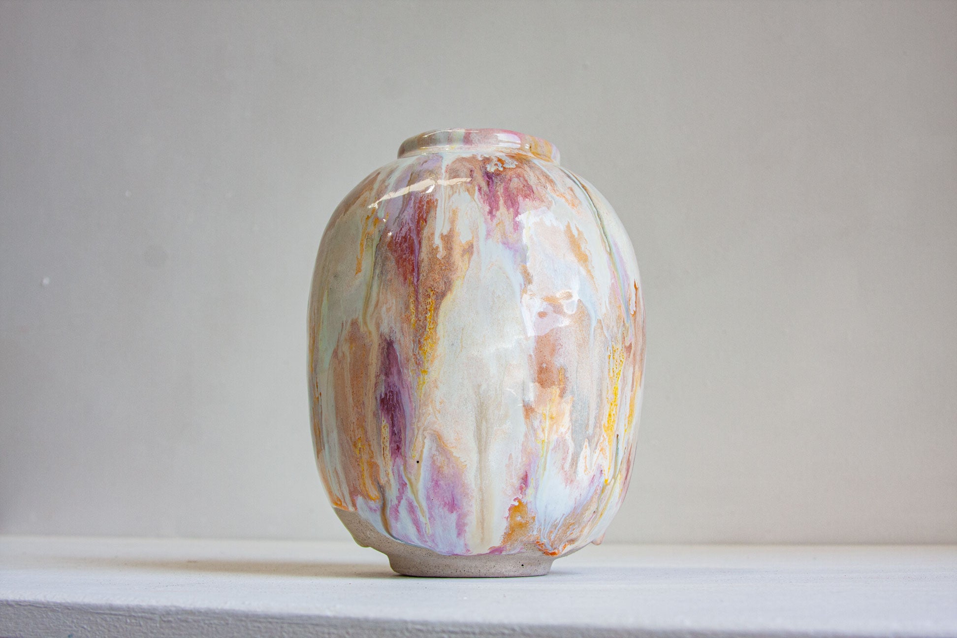 A small ceramic vase with a glossy finish, featuring a marbled pattern of white, purple, orange, and gold, set against a plain light gray background.