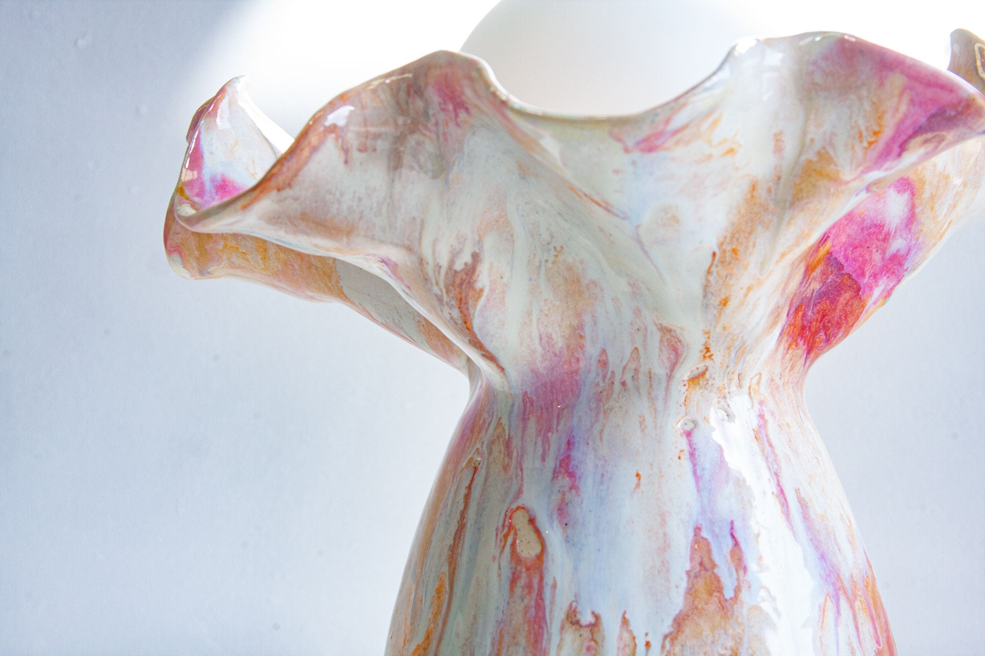A close-up of a sculptural ceramic vase with a flared, organic rim, decorated in a marbled glaze of white, pink, peach, and orange on a light gray background.