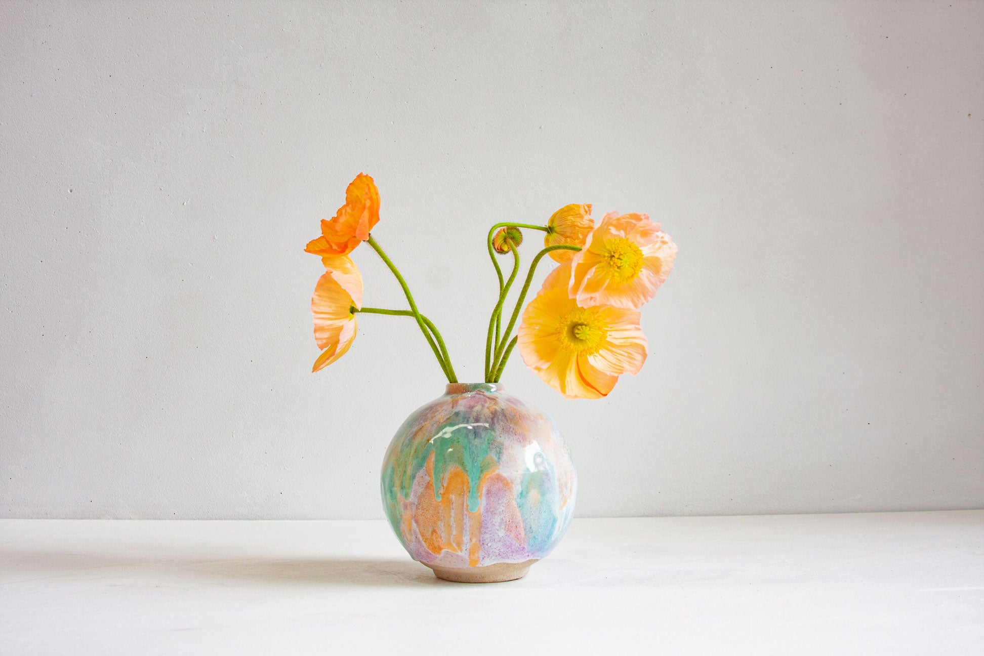 A round handmade ceramic vase glazed in pastel purple, peach and green, holding orange and yellow poppies against a plain pale background.