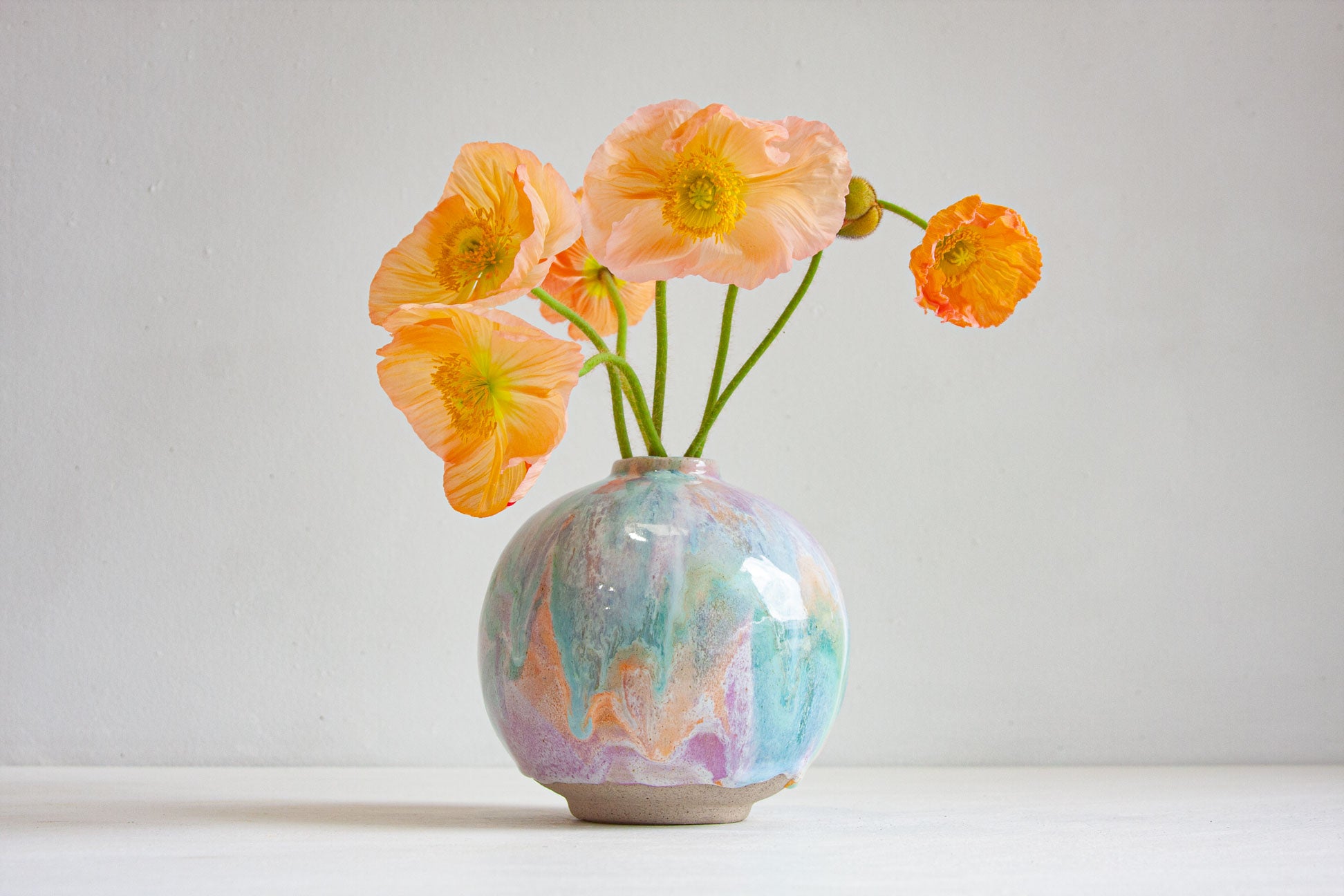 A round handmade ceramic vase glazed in pastel purple, peach and green, holding a loose arrangement of peach poppies.