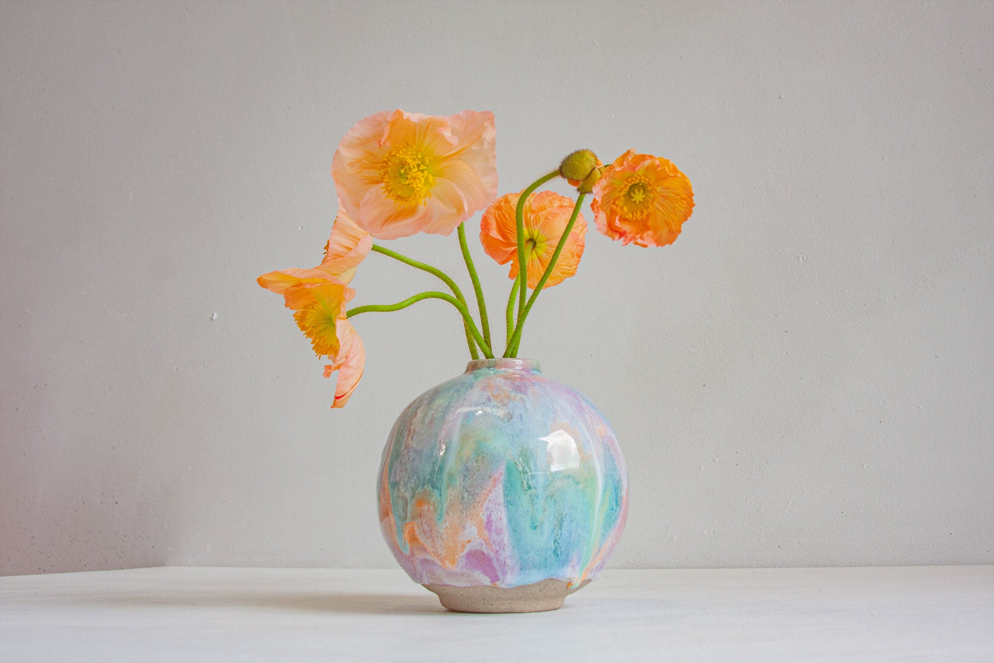 A round handmade ceramic vase glazed in pastel purple, peach and green, holding peach and orange poppies, against a plain pale wall.