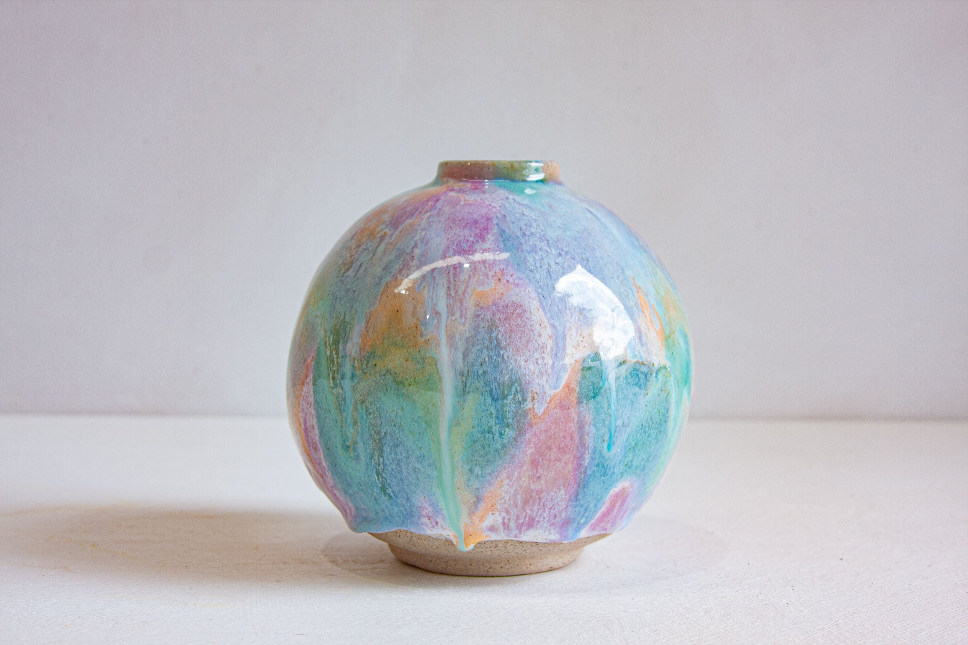 A round handmade ceramic vase photographed front on, the pastel purple, peach and green glaze running down to a bare speckled clay foot.