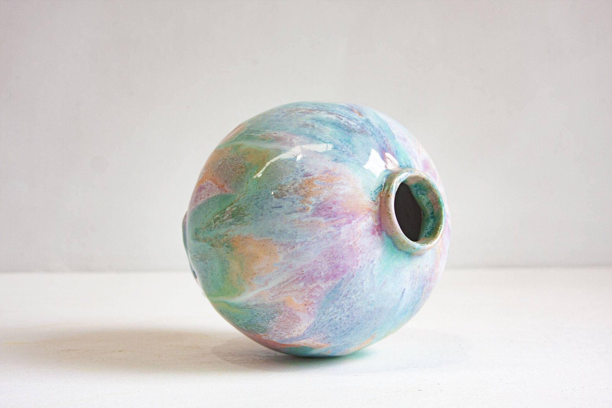 A round handmade ceramic vase tipped to show its small neck opening, glazed in drifting pastel purple, peach and green.