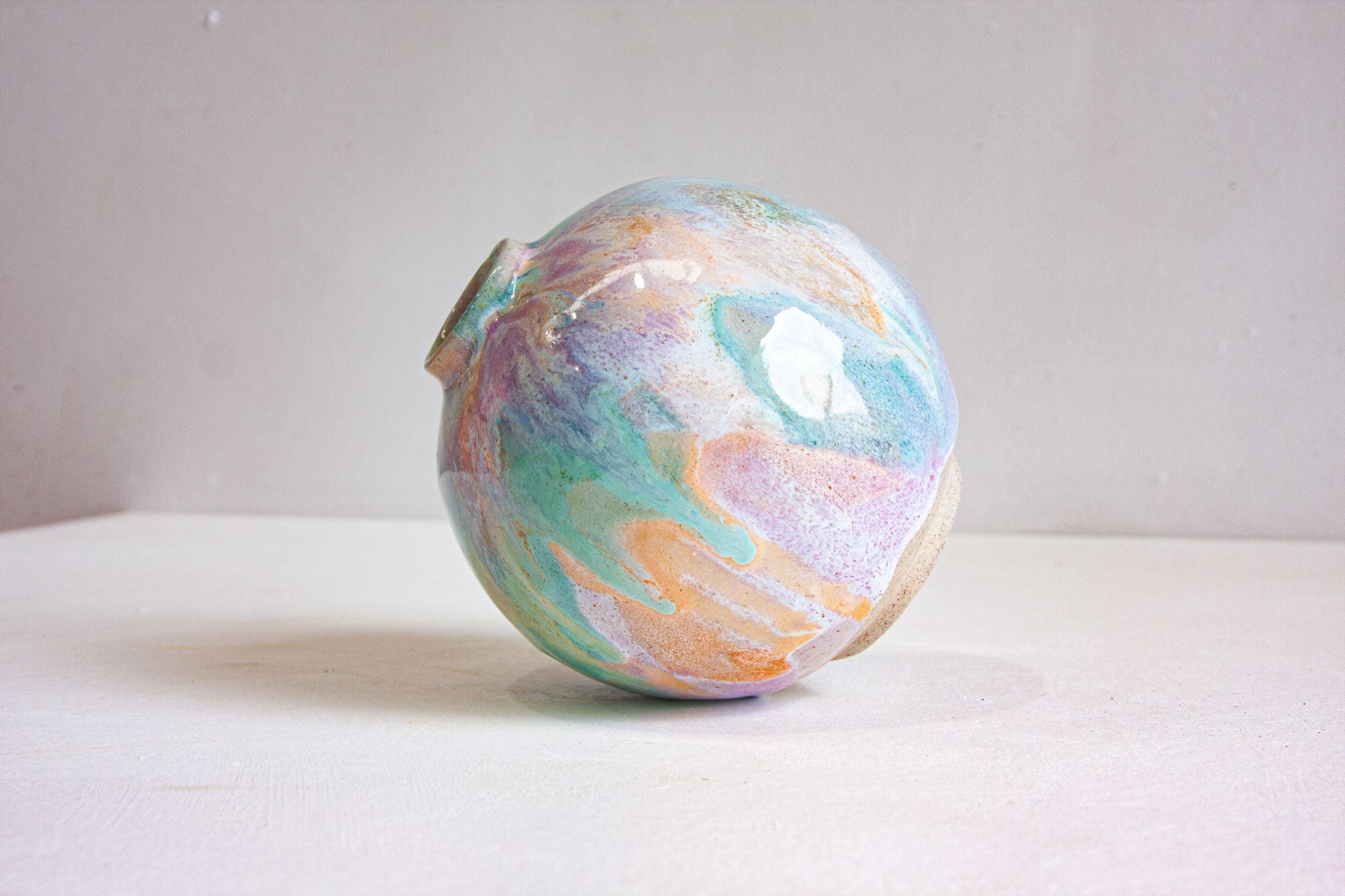A round handmade ceramic vase seen from a low angle, the pastel purple, peach and green glaze curving over the shoulder of the form.