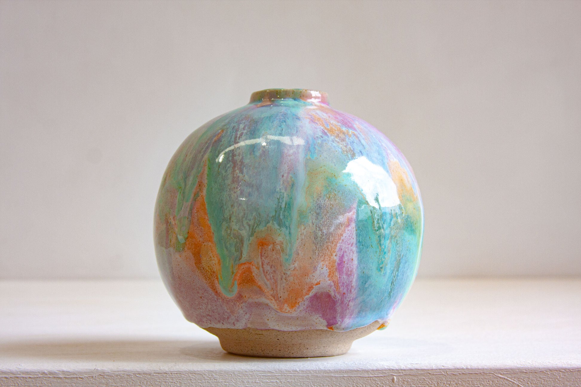 A round handmade ceramic vase front on, pastel purple, peach and green falling in soft vertical drifts over the globe form.