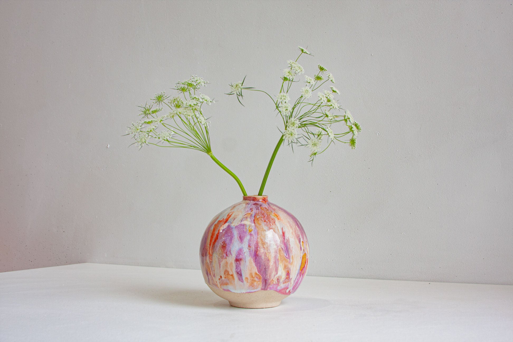 A round ceramic vase with a marbled pink, purple, and orange pattern, holding two green stems with small white flowers, set against a neutral light grey background.