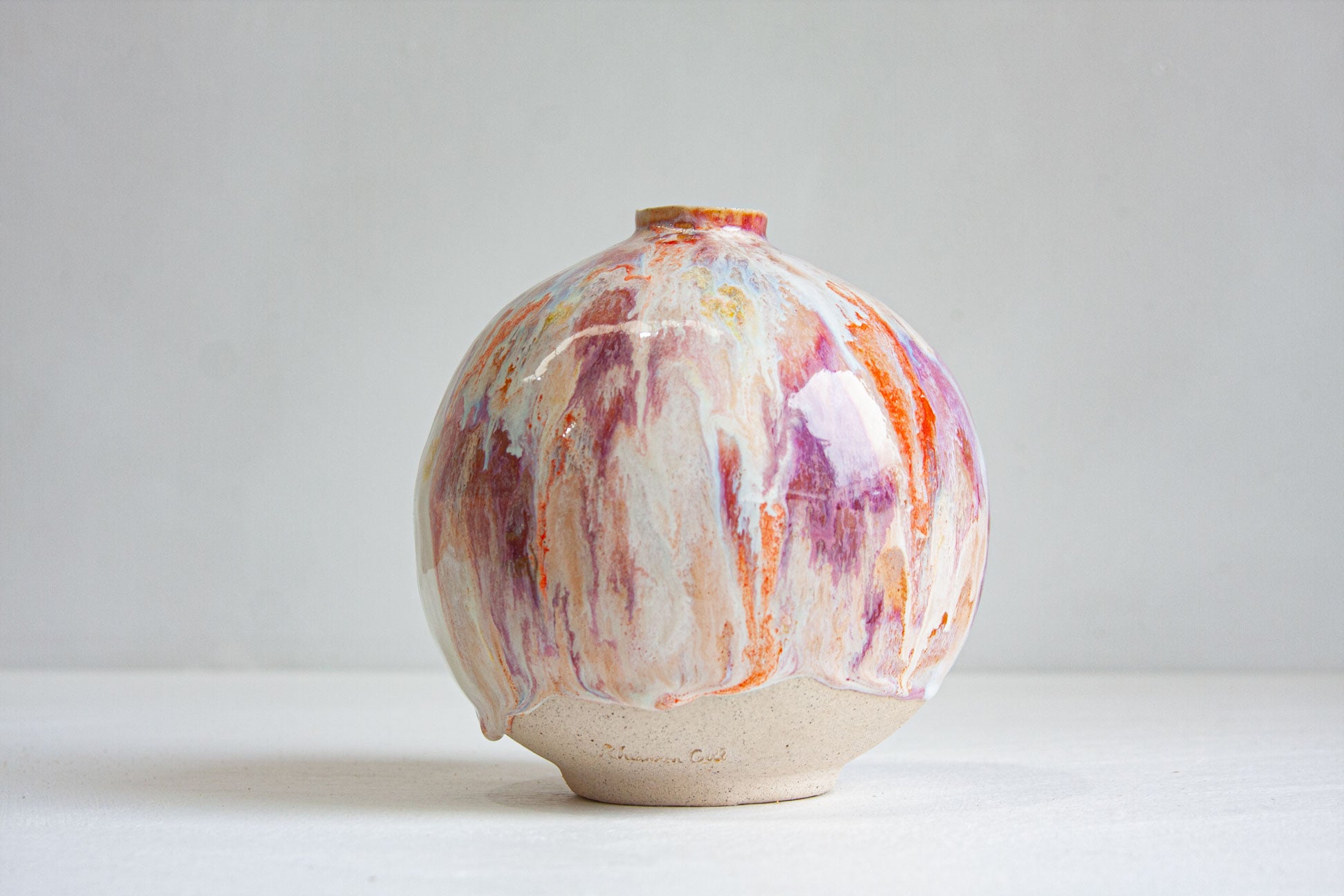 A handmade ceramic vase with a rounded, bulbous shape, featuring a marbled glaze pattern of orange, purple, pink, and cream on a light beige base, set against a plain light grey background.