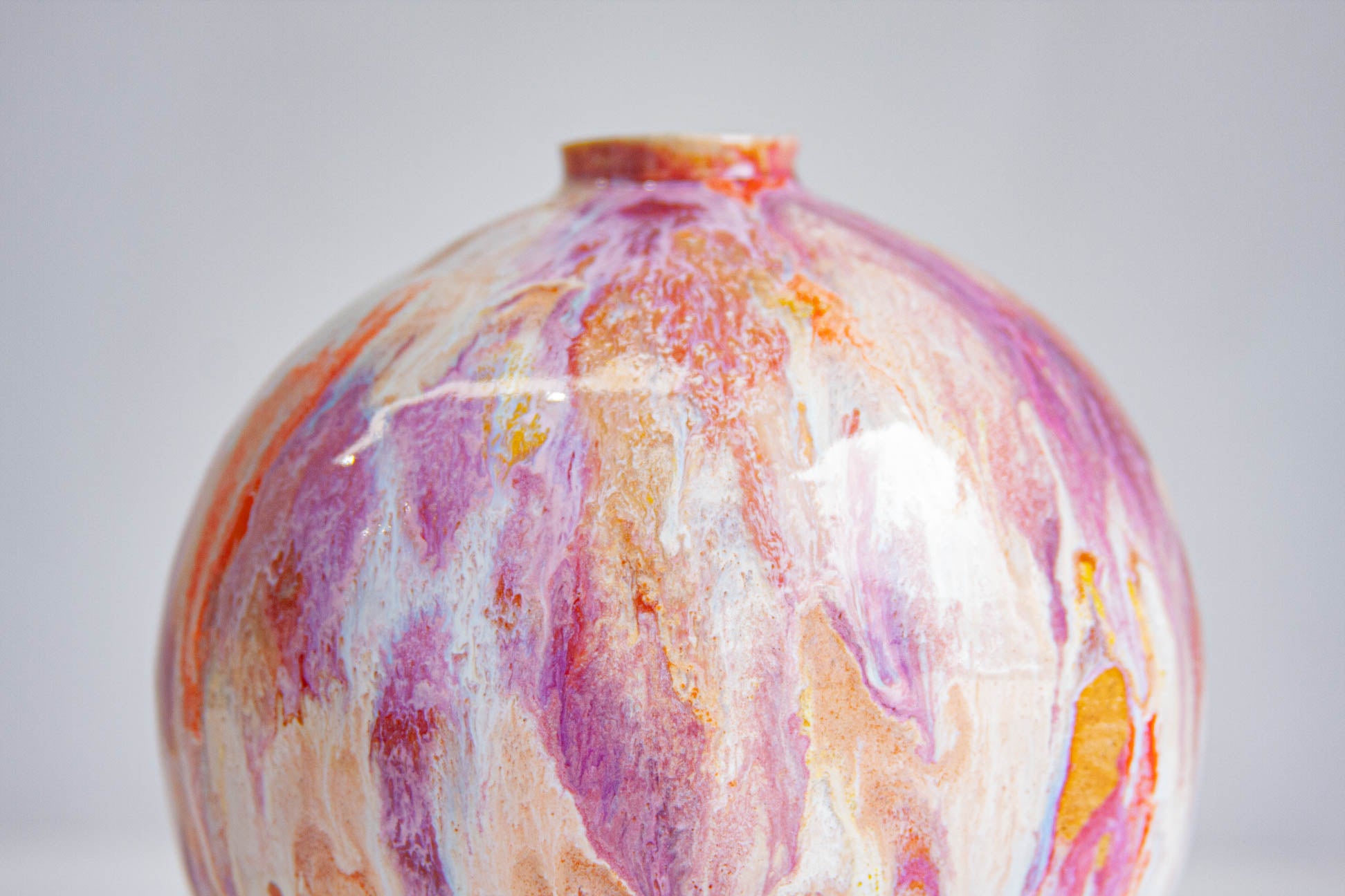 A close-up of a round ceramic vase with a glossy finish, featuring a marbled pattern of pink, orange, white, and purple glaze on a light grey background.