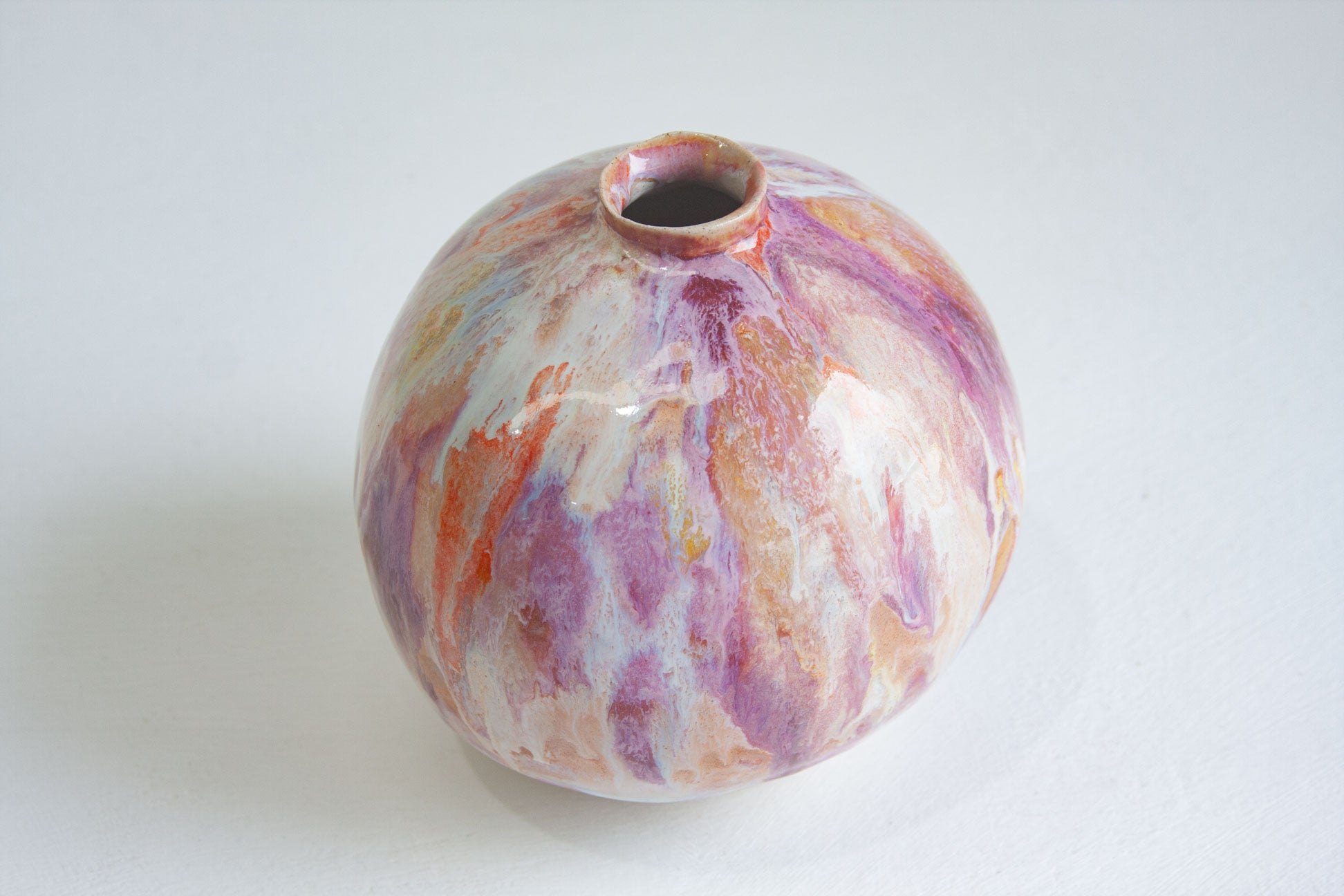 A small round ceramic vase with a narrow neck and open top, decorated with a marbled pattern of purple, orange, pink, and white glaze on a light grey background.