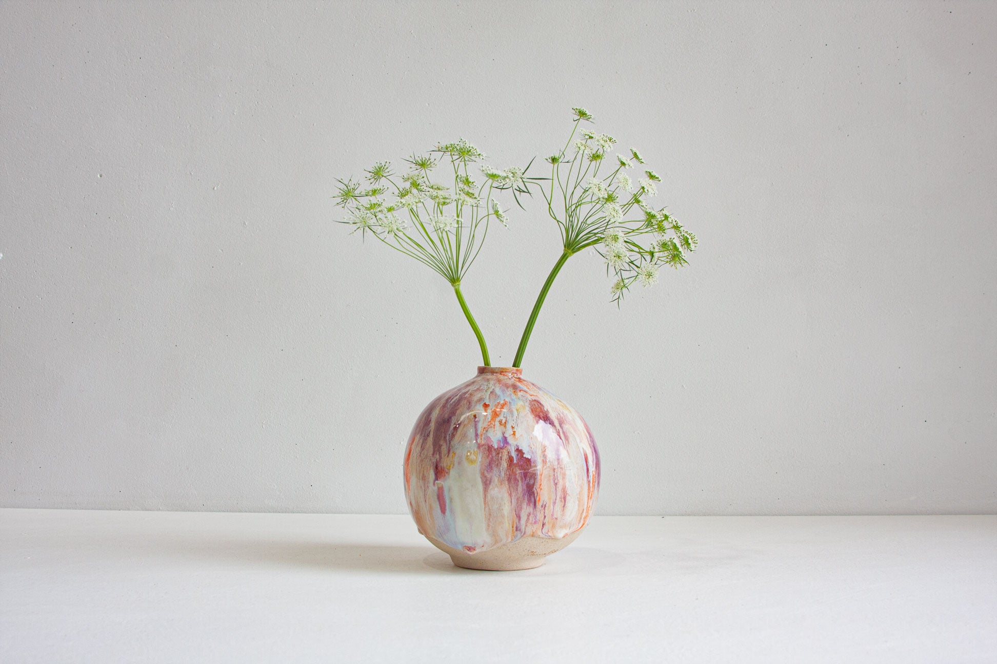 A small round ceramic vase with a marbled pink, purple, and cream glaze, holding two green stems of delicate white umbellifer flowers, set against a plain light grey background.