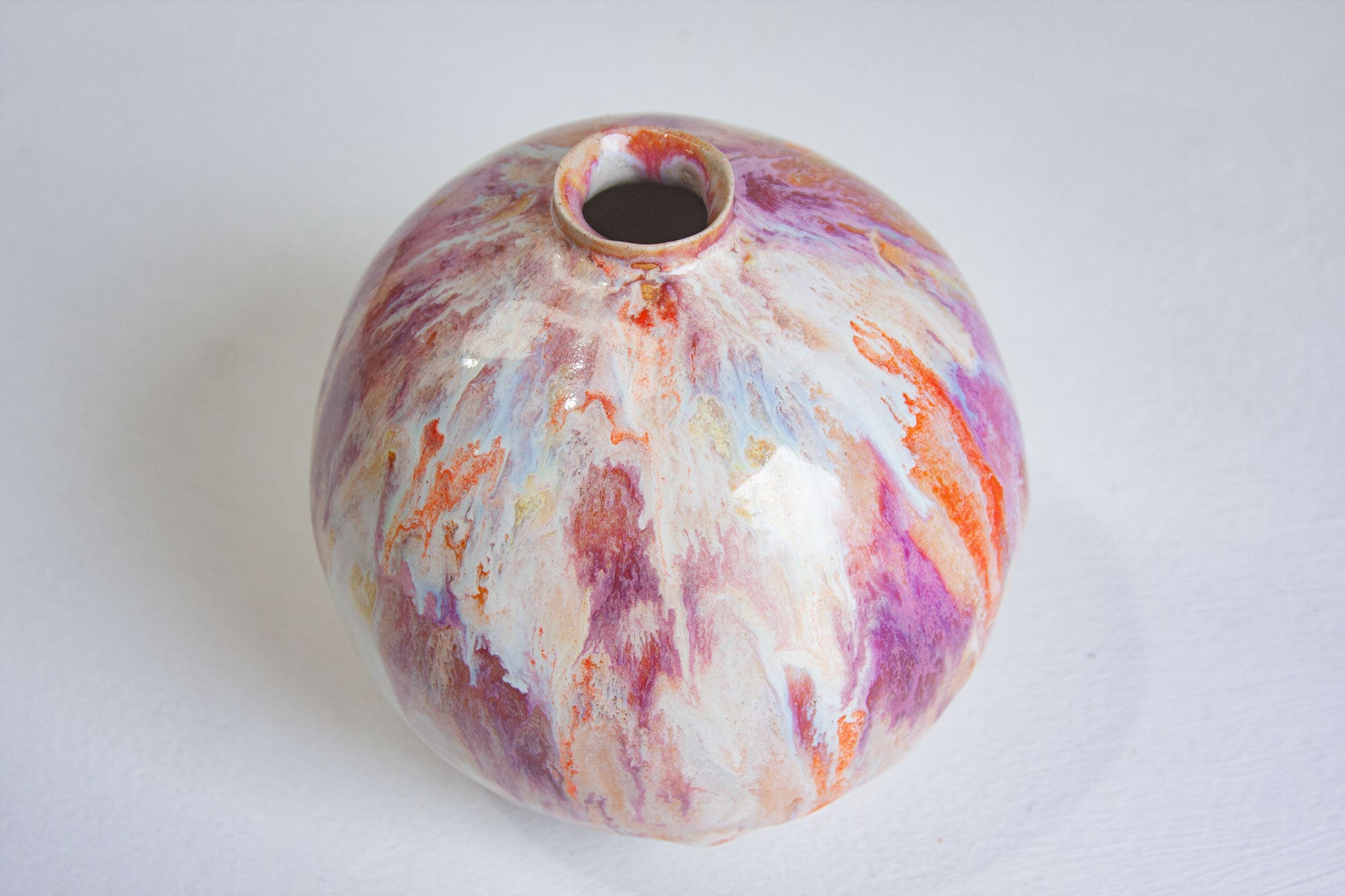 A small round ceramic vase with a glossy finish, featuring a marbled pattern of purple, orange, pink, and white on a white background.
