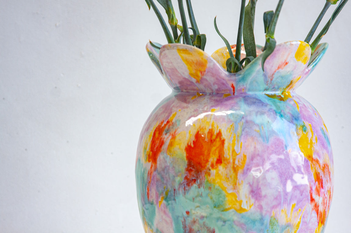 Close-up of the ruffled petal-shaped rim of handmade ceramic sculptural colour theory vase showing orange, yellow, teal and lavender multicolour glaze detail, with carnation stems