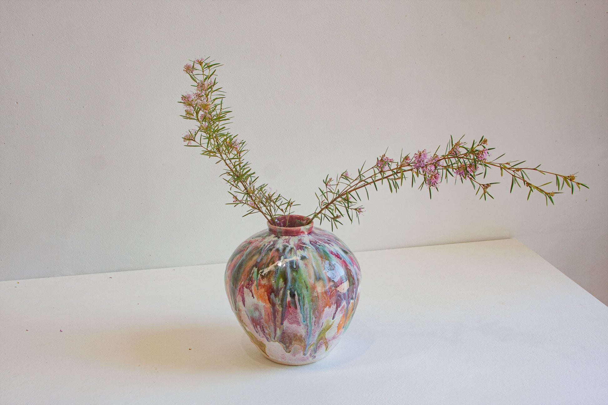 A small round ceramic vase with a colorful marbled pattern in pink, green, orange, and white, holding a delicate flowering green branch against a plain off-white background.