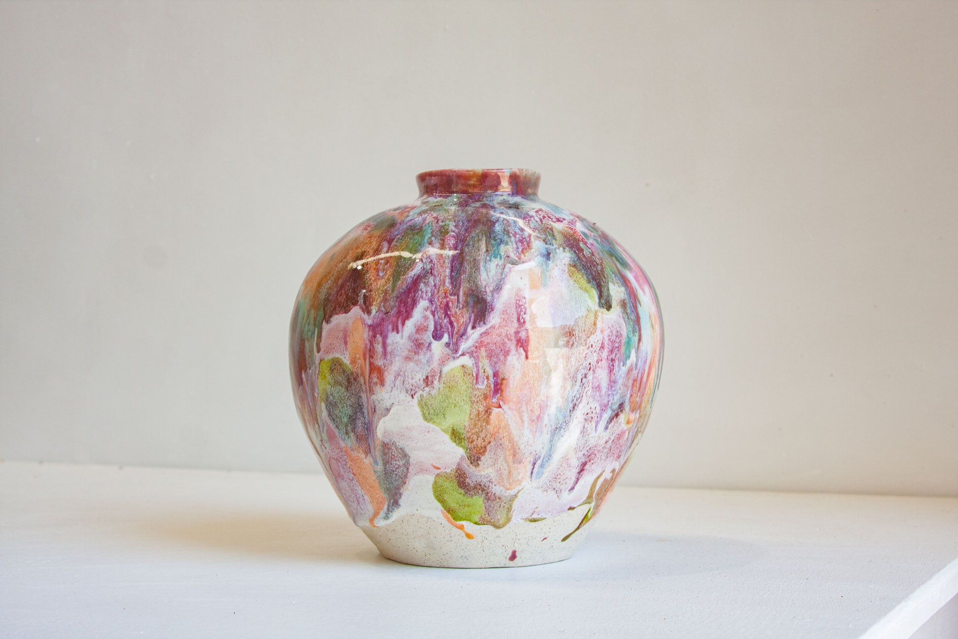 A small ceramic vase with a rounded body and narrow neck, decorated with a marbled, fluid-art pattern in pink, purple, green, orange, and white, sitting on a white surface against a light beige wall.
