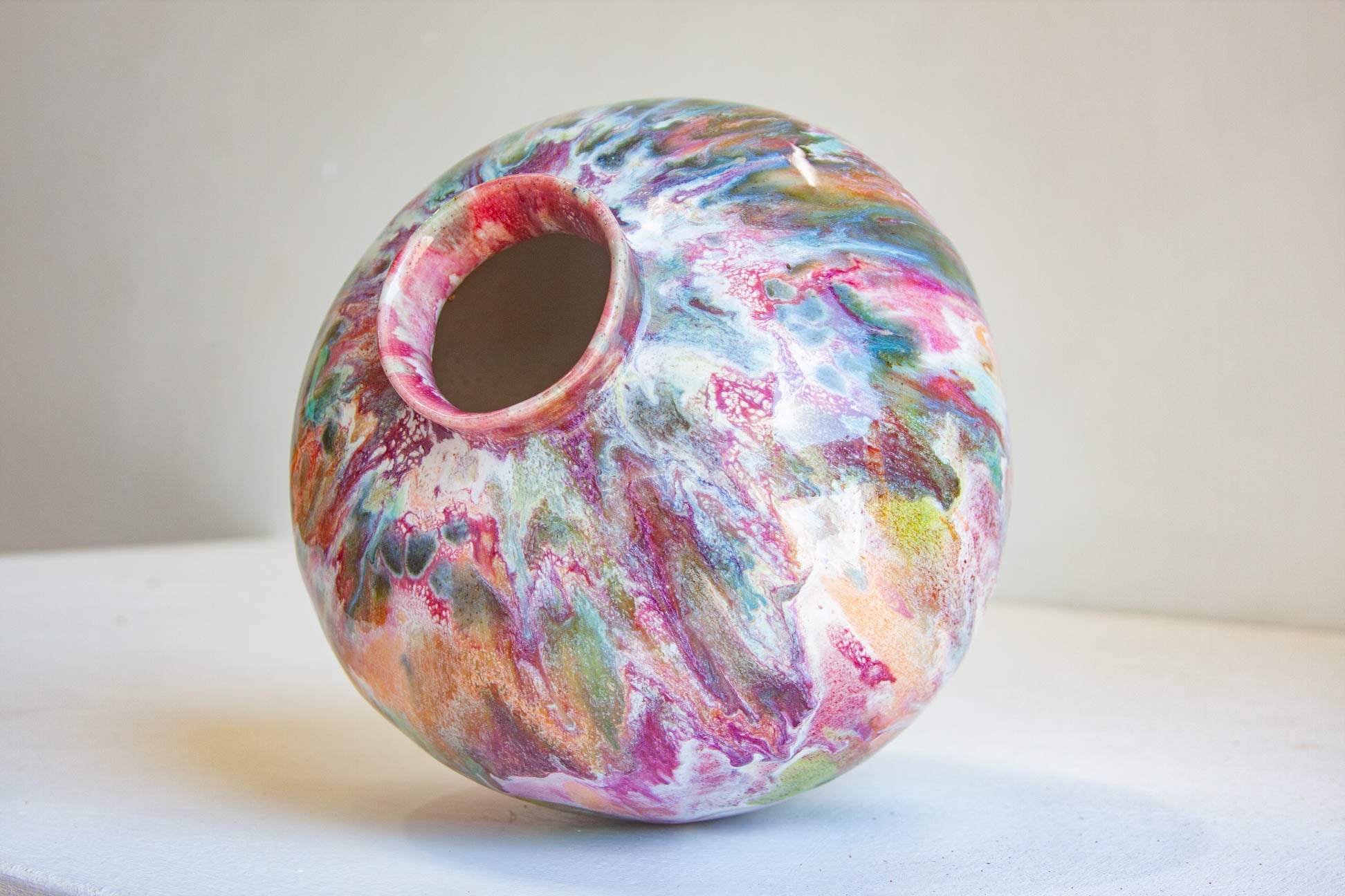 A round ceramic vase with a colorful marbled pattern in pink, purple, blue, green, and white, sitting on a light surface against a neutral background.