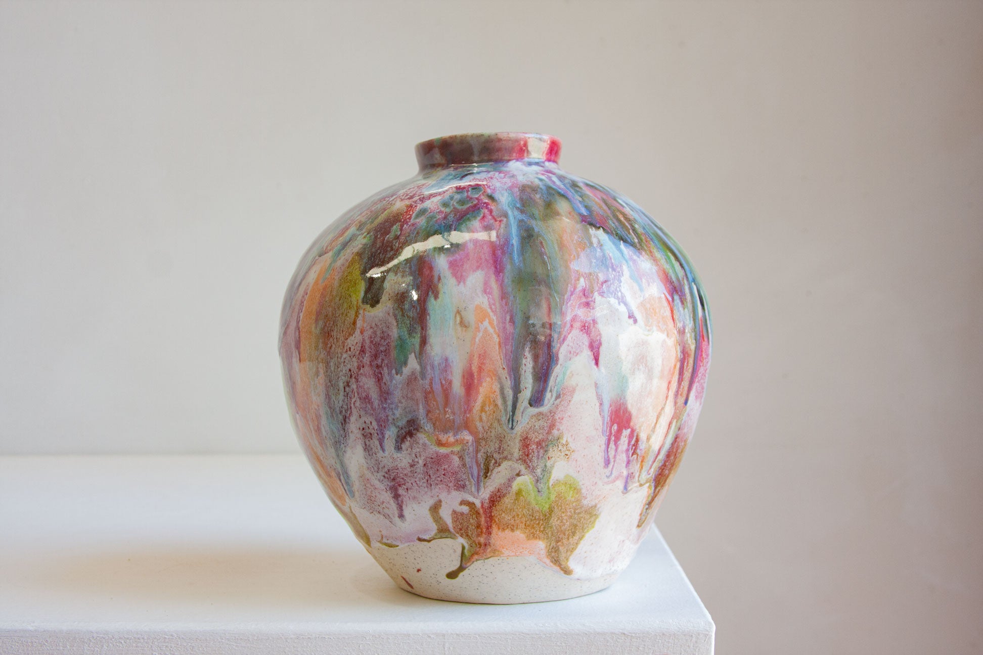 A round ceramic vase with a marbled, abstract pattern in pink, purple, blue, green, and gold on a white base, displayed on a white pedestal against a neutral beige background.