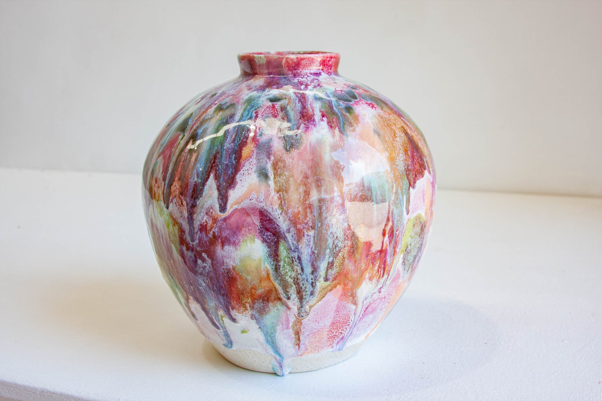 A small ceramic vase with a rounded body and short neck, decorated with a colorful abstract marbled glaze in pink, purple, blue, green, and orange tones, set against a plain light background.