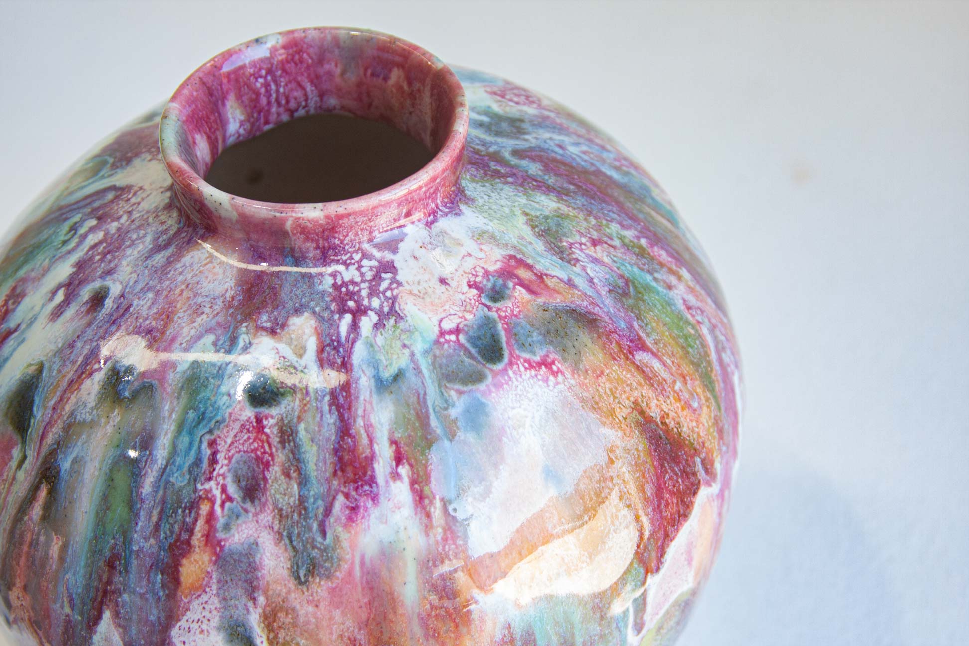 A close-up of a ceramic vase with a rounded body and short flared rim, covered in a marbled glaze with swirling pink, purple, blue, green, and white tones on a light gray background.