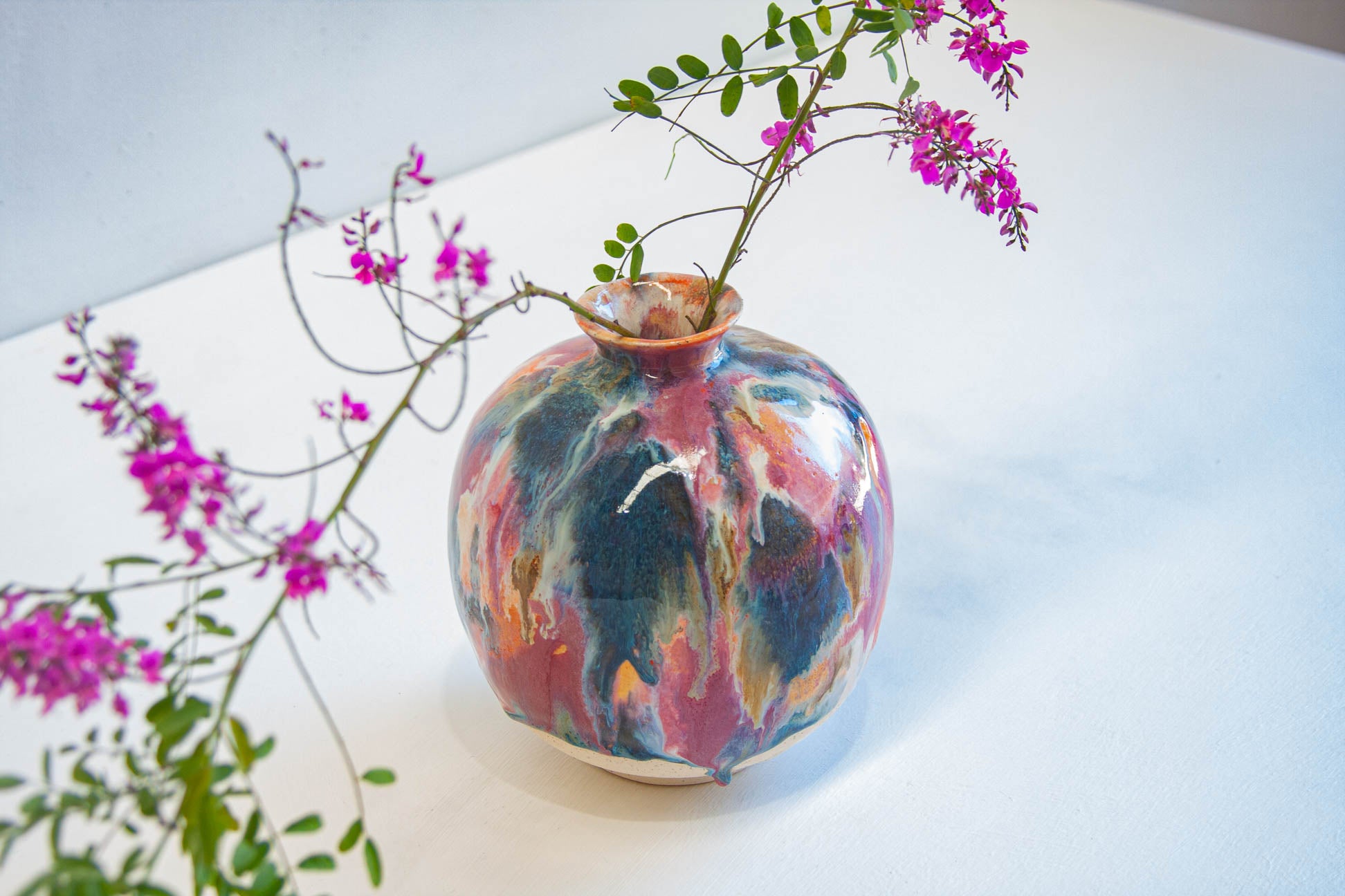 A round ceramic vase with a marbled, swirled glaze in blue, pink, orange, and white, holding delicate pink flowers on a white surface.