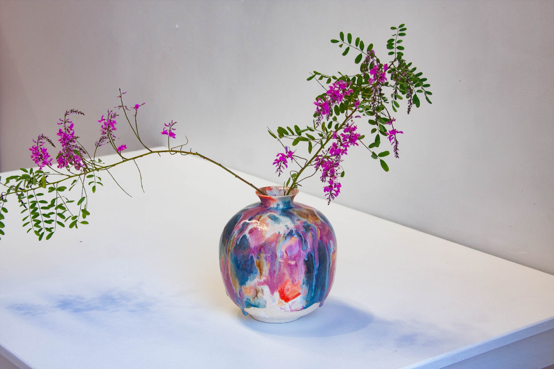 A colorful marbled ceramic vase with pink, blue, purple, and orange tones, holding a flowering branch with green leaves and small pink blossoms, set on a white surface against a light gray wall.