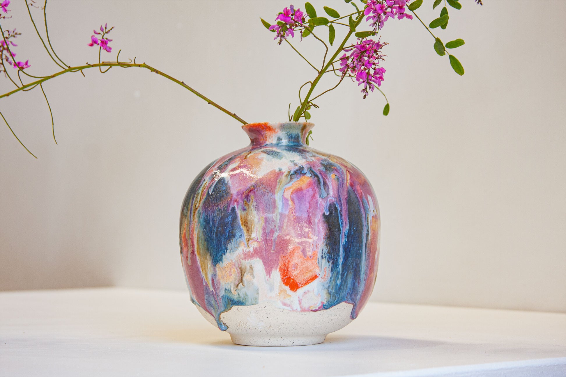 A ceramic vase with a colorful, dripping glaze pattern in shades of blue, pink, purple, and orange, holding a flowering branch with pink blossoms against a neutral cream background.