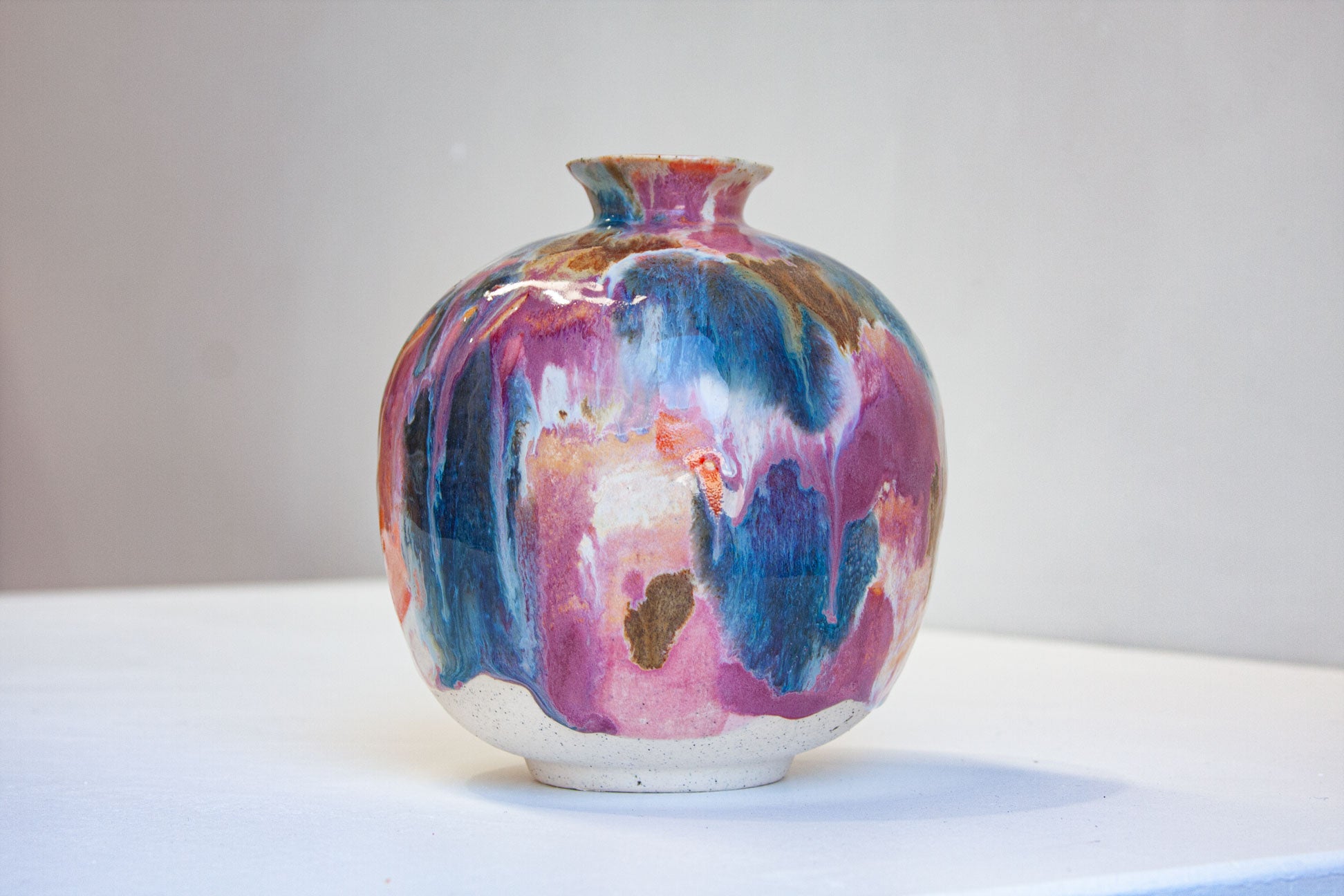 A small round ceramic vase with a narrow neck, decorated in a marbled, drip-style glaze of pink, blue, purple, white, and brown tones, set against a plain light gray background.