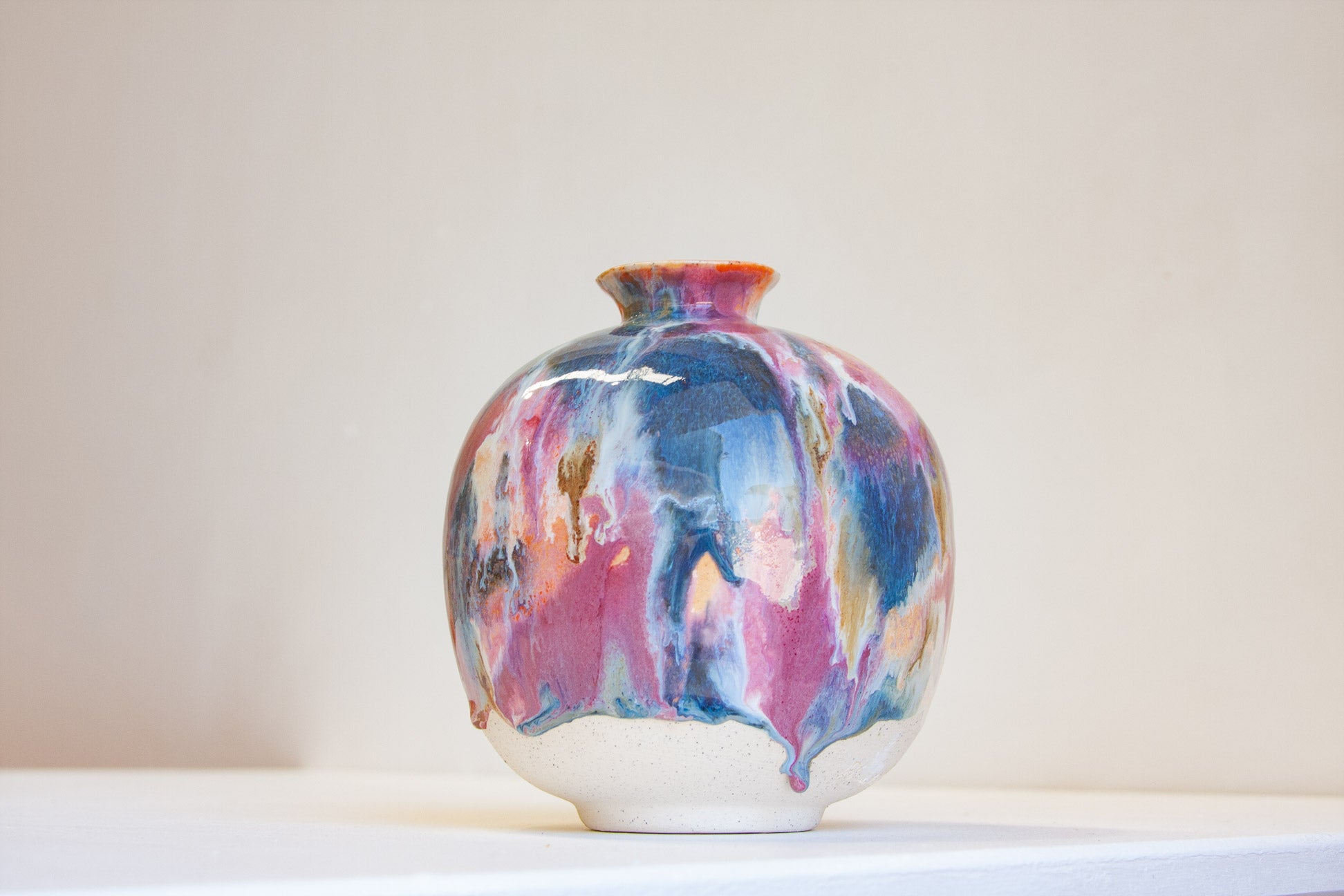 A small ceramic vase with a rounded body and short flared neck, decorated with a marbled, drip-style glaze in blue, pink, purple, and orange over a white base, set against a plain light beige background.