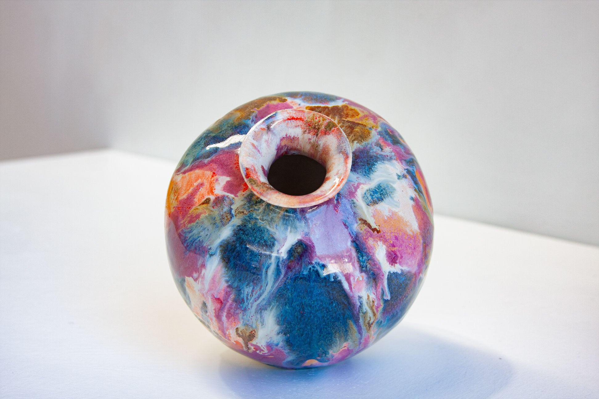 A round ceramic vase with a marbled, abstract pattern of blue, pink, purple, orange, and white, featuring a small flared opening at the top, set against a plain light gray background.