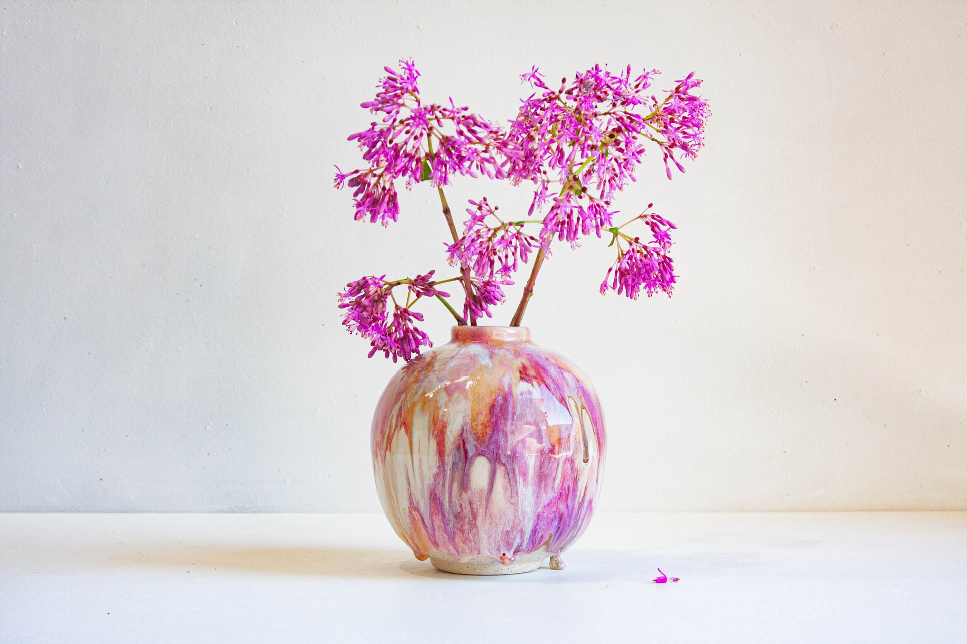 A round ceramic vase with a marbled pink, purple, and white glaze, holding pink-purple flowers against a plain off-white background.