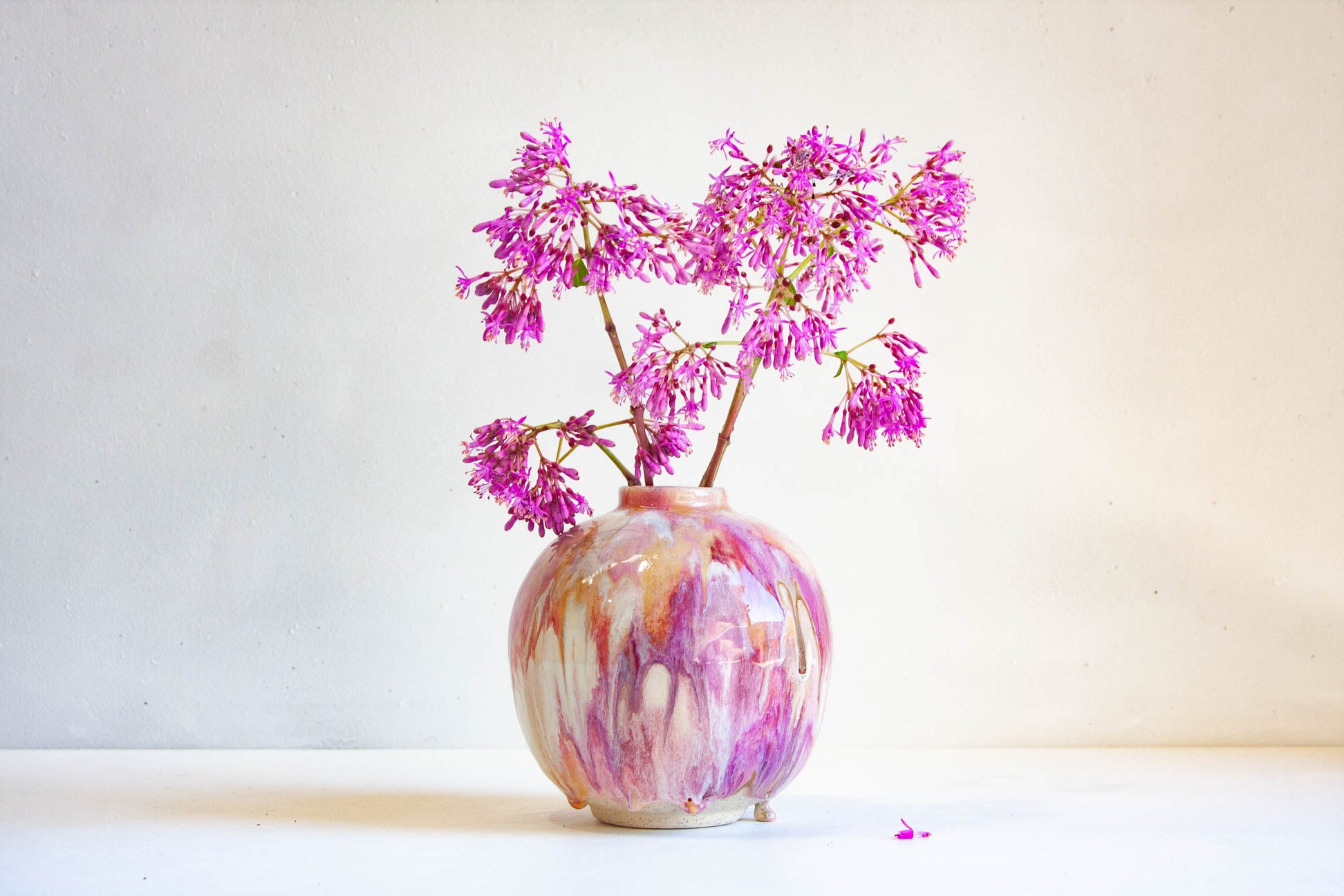 A round ceramic vase with a marbled pink, purple, and cream glaze, holding delicate pink-purple flowering branches against a plain off-white background.